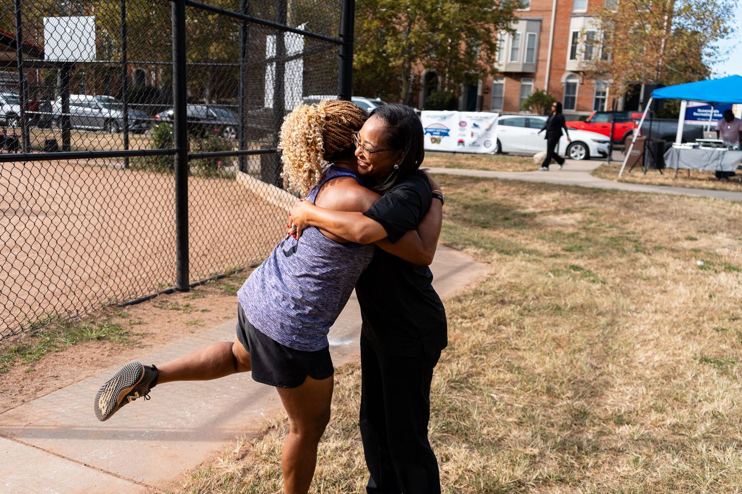 Two women share a warm embrace outdoors near a chain-link fence on a sunny day.