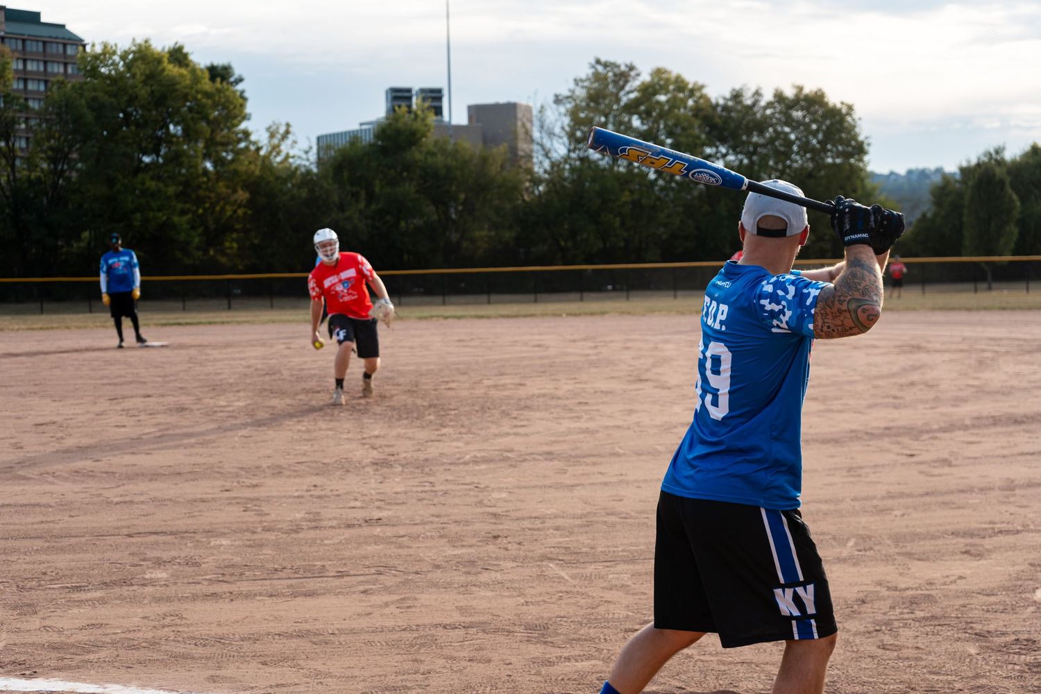 Players compete in a recreational softball game on a dirt field, with a batter in blue jersey preparing to swing.