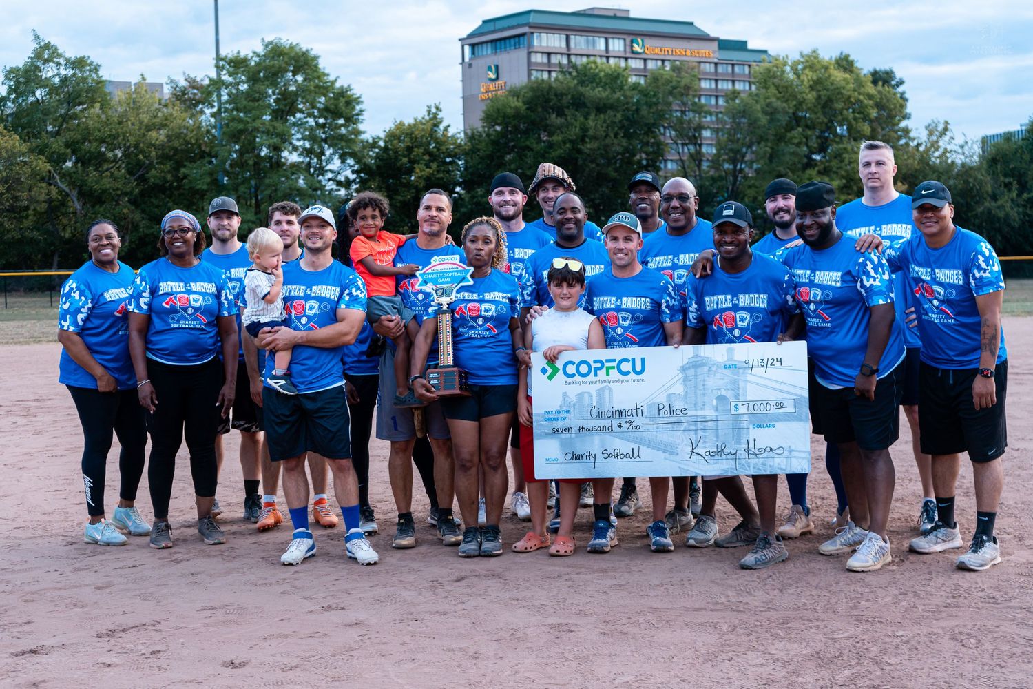 Group of people in blue COPFCU shirts pose on softball field holding large charity check outdoors.