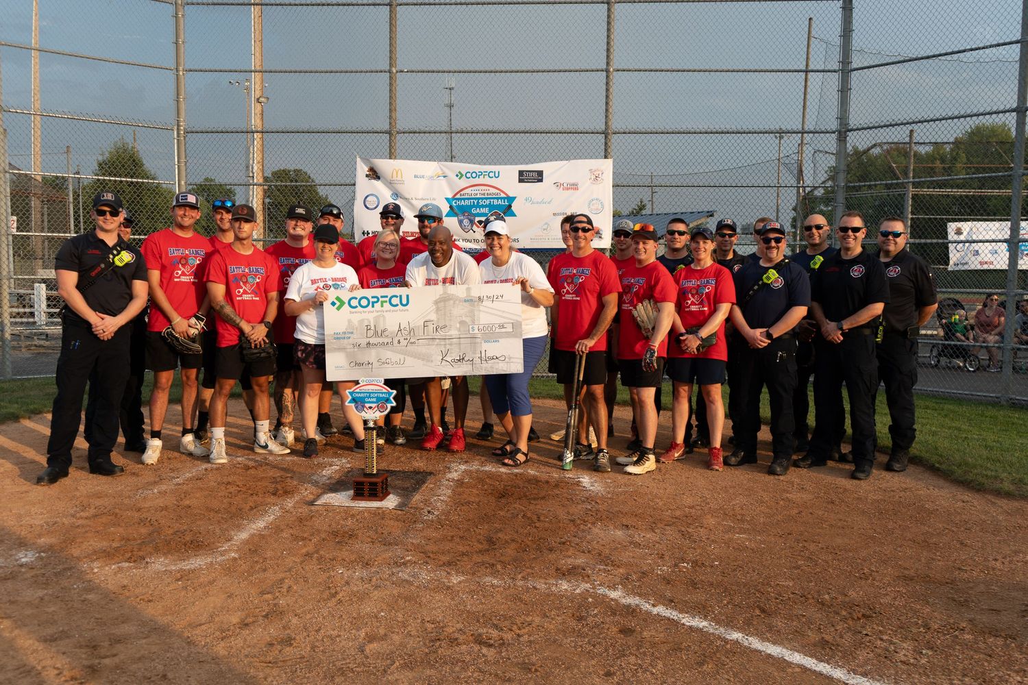 Softball team celebrates championship win holding large COPCU check at home plate with trophy and supporters.