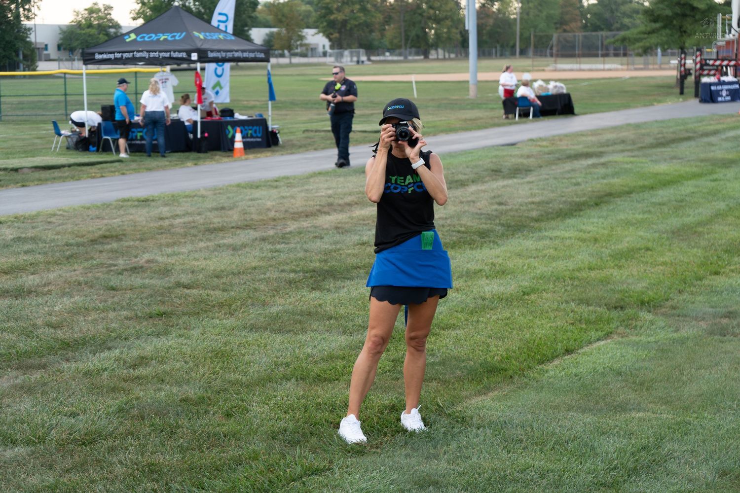 Woman in blue skirt photographs outdoor event on green field with tent and people in background.