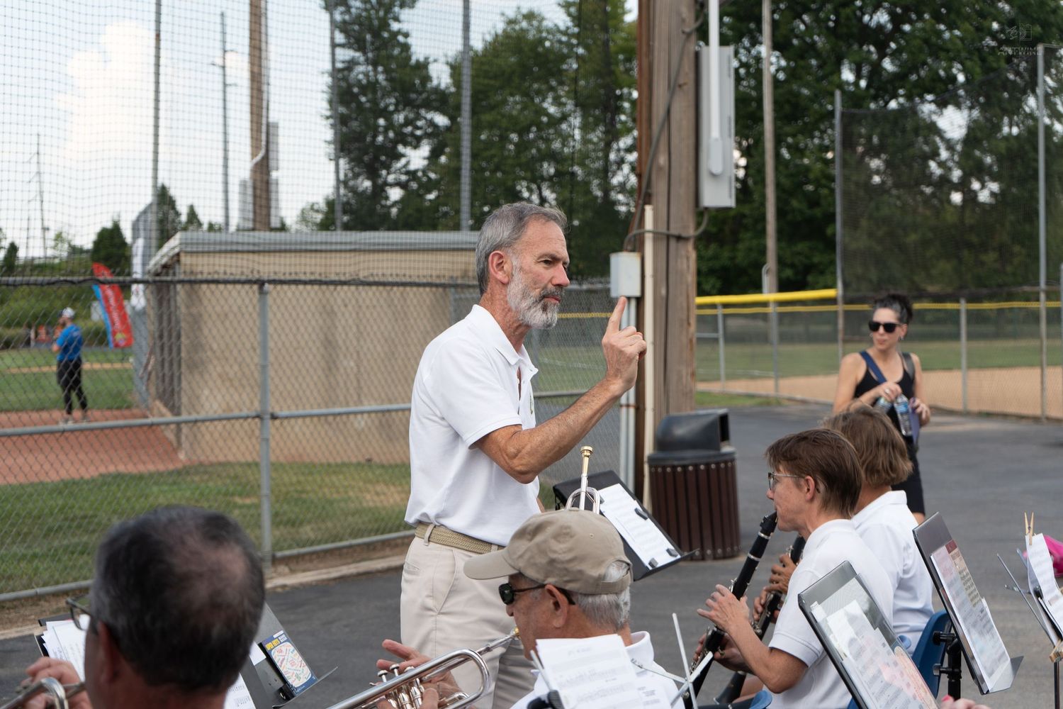 Outdoor band conductor directing musicians at a community concert near a baseball field on a sunny day.