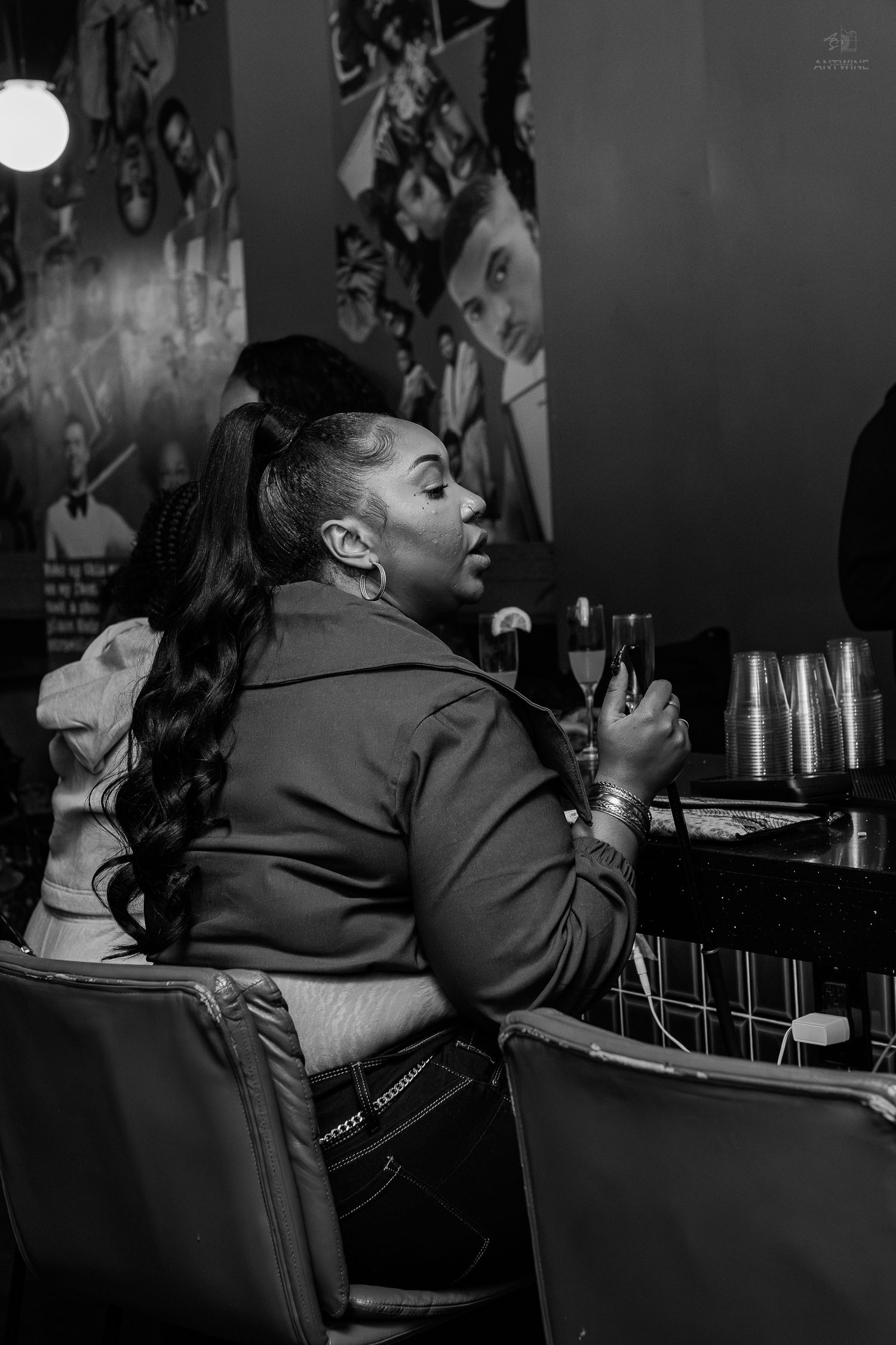 Black and white photo of a woman sitting at a bar, holding a drink glass in a dimly lit venue.