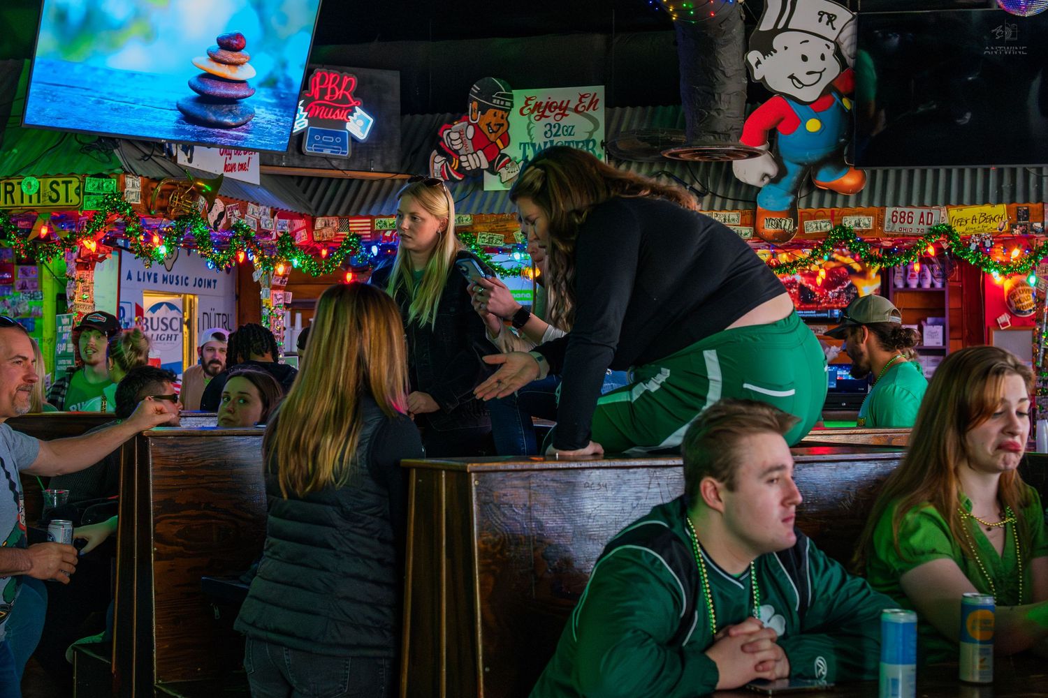 Lively bar scene with patrons in green outfits celebrating St. Patrick's Day amid colorful decorations.