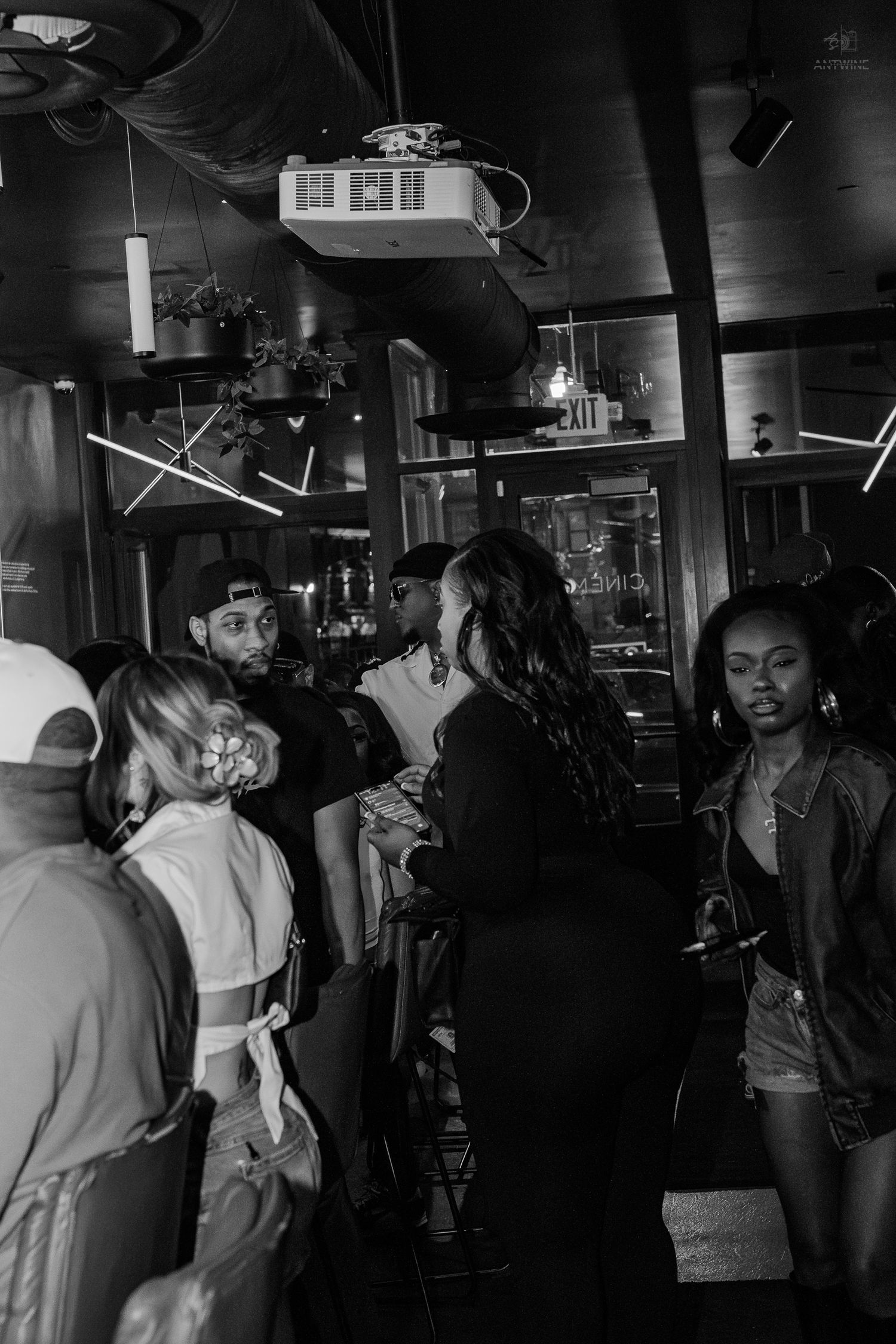 Black and white photo of a diverse group of people socializing inside a dimly lit bar or lounge.