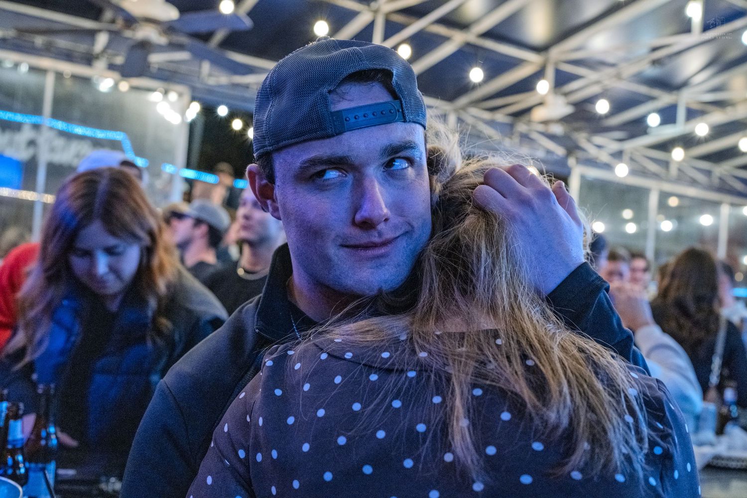 Young man in a backwards cap smiling at a crowded indoor event venue.