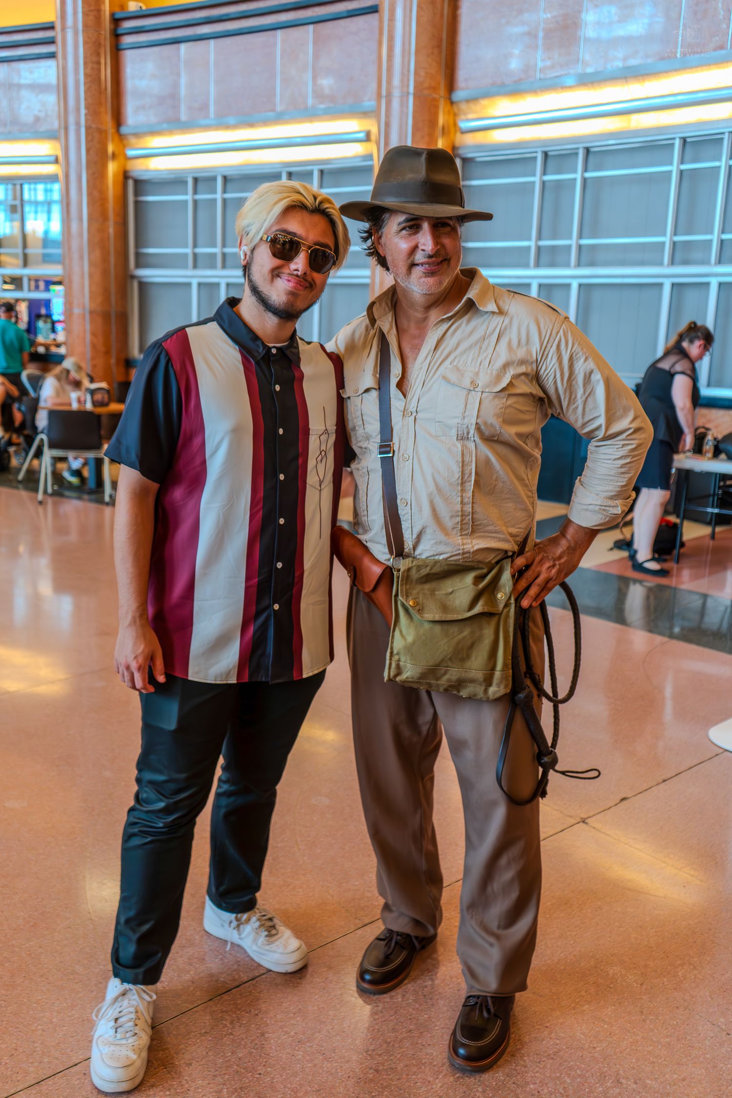 Two men posing together at an indoor event, one dressed as Indiana Jones in costume with hat and satchel.