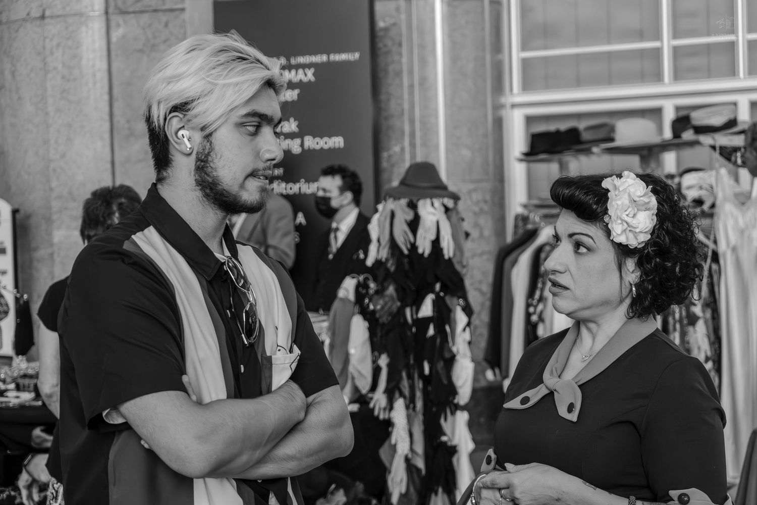 Black and white photo of two people in vintage attire conversing at an indoor market event.