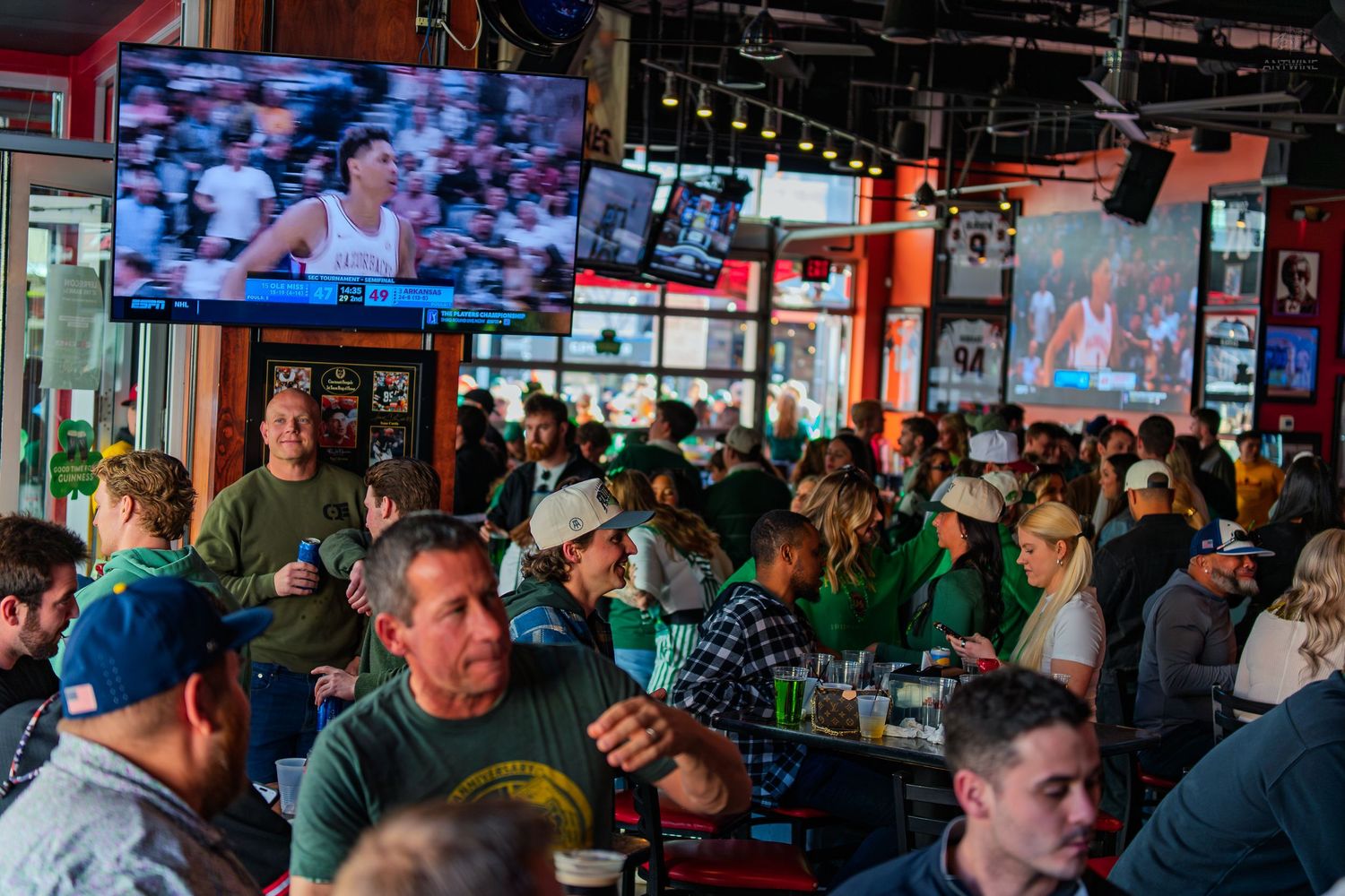 Crowded sports bar with fans watching basketball on TVs, wearing green shirts, enjoying drinks at tables.