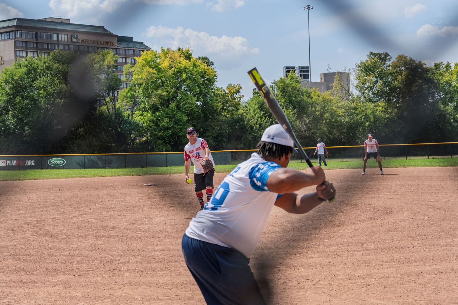 Softball batter prepares to swing at pitch during outdoor charity softball game on sunny day.