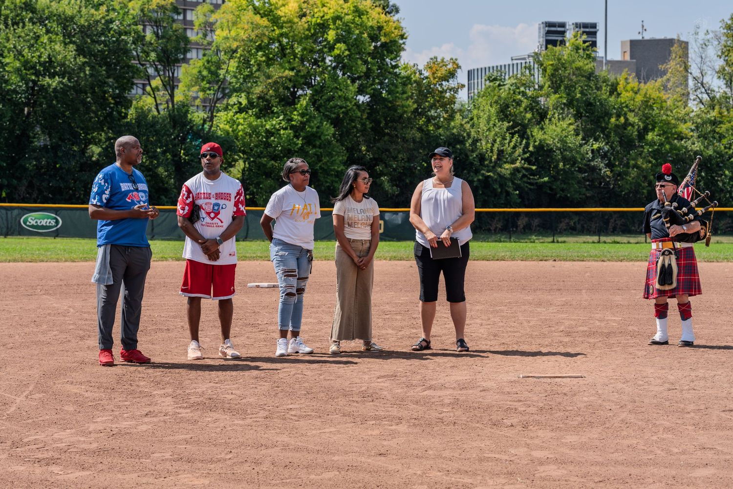 Group of people standing on a baseball diamond during an outdoor ceremony with a bagpiper.