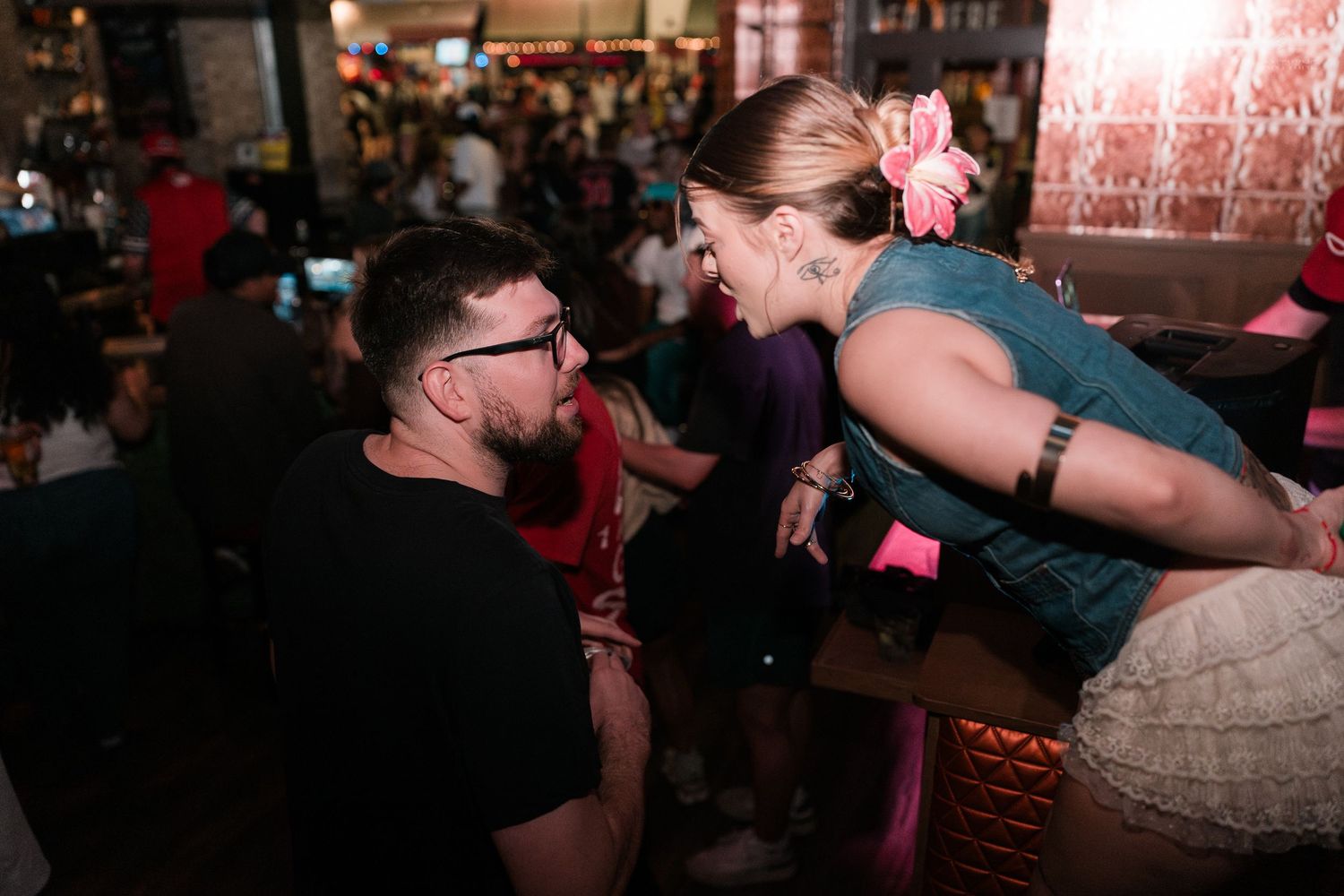 Man in black shirt and woman in denim vest laughing together closely at a busy bar.