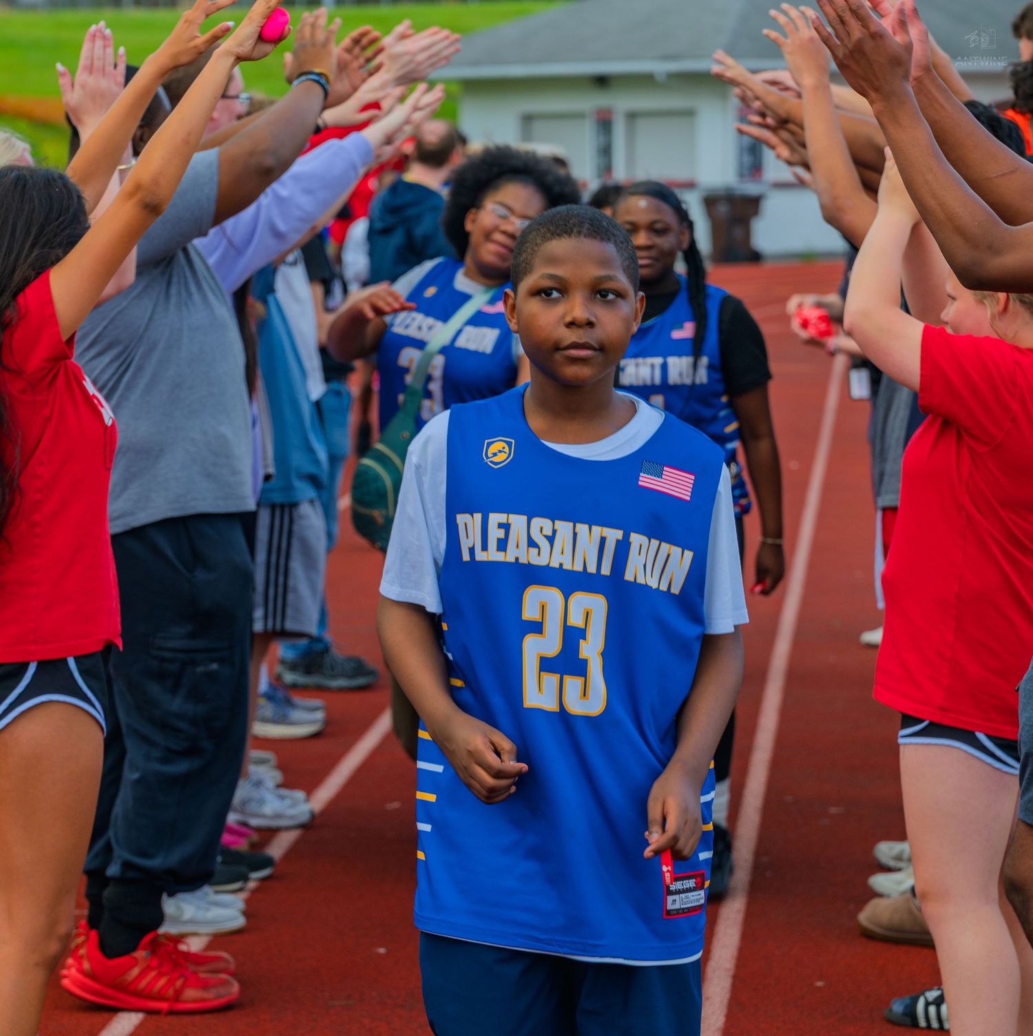 Young athlete in Pleasant Run #23 blue jersey walks through a celebratory tunnel of raised hands on a track.