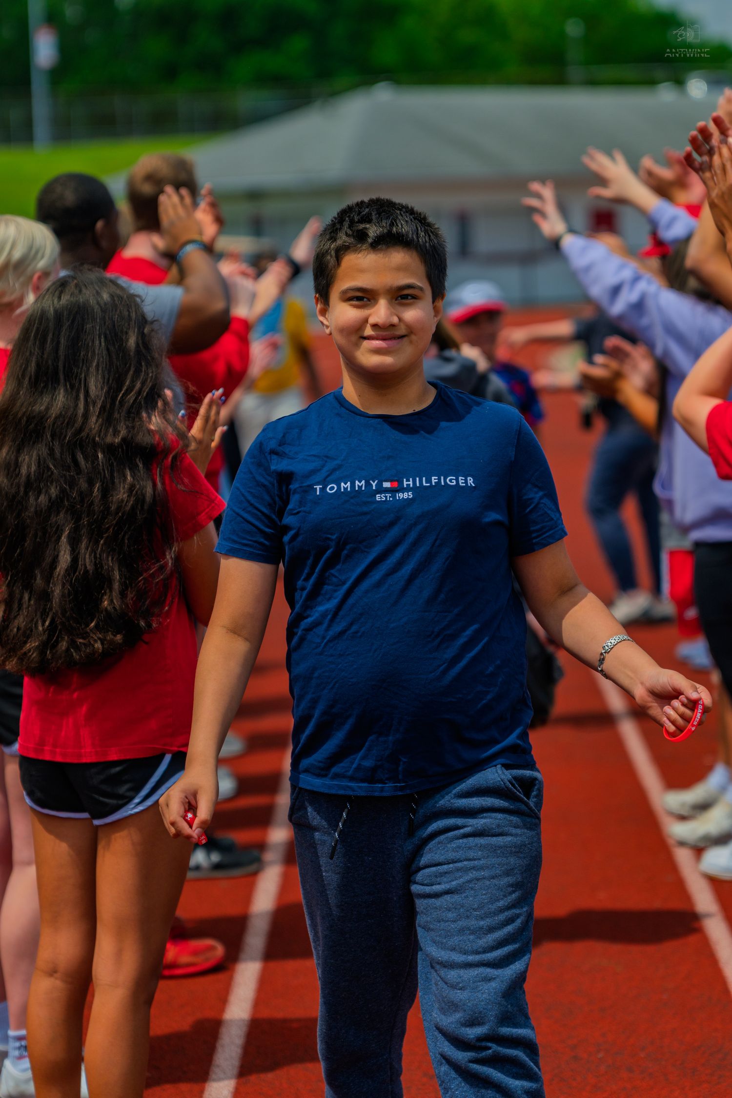 Young student in Tommy Hilfiger shirt walks track at school event while peers high-five around him.