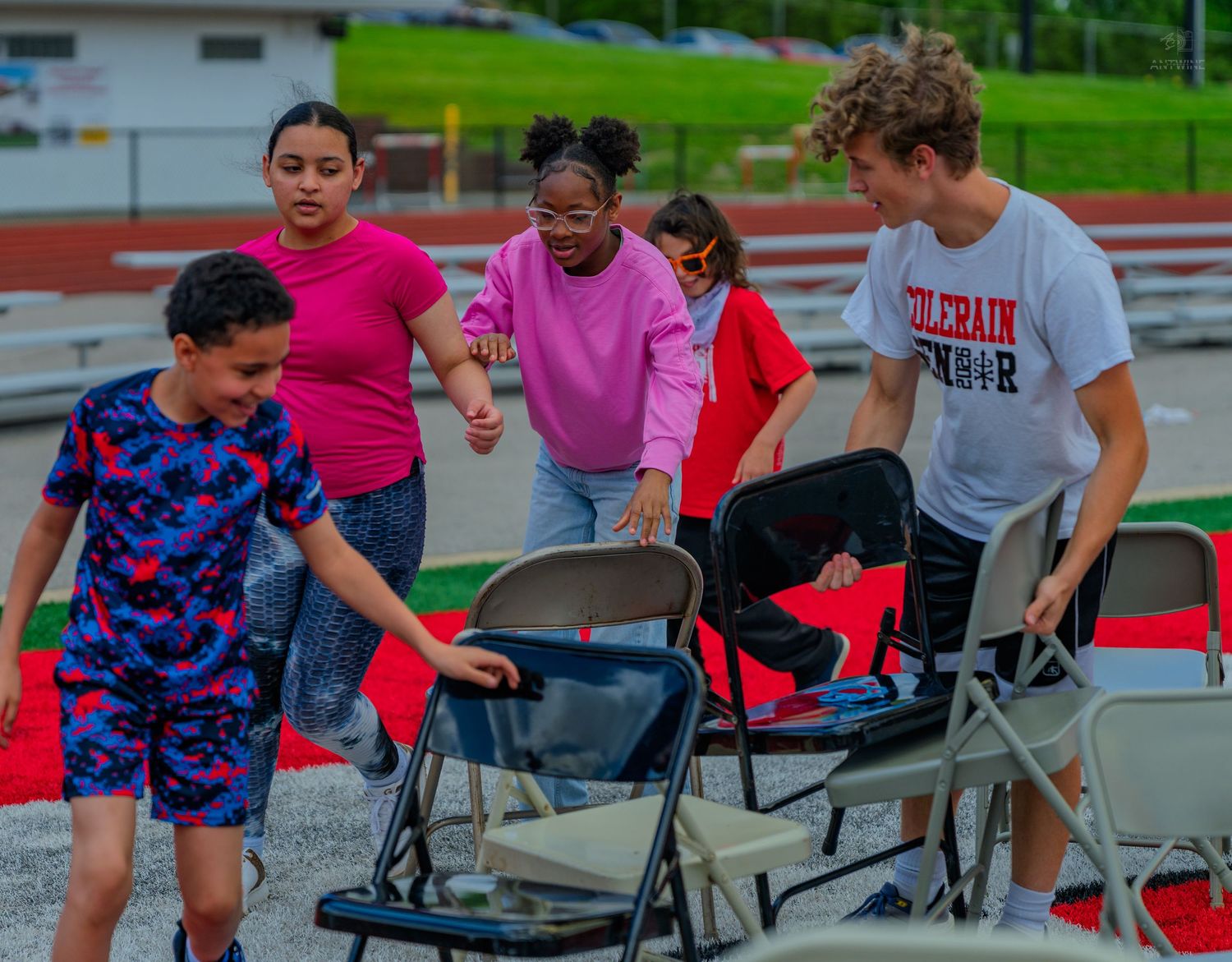 Children and a teen playing musical chairs outdoors on a track during a fun school activity event.