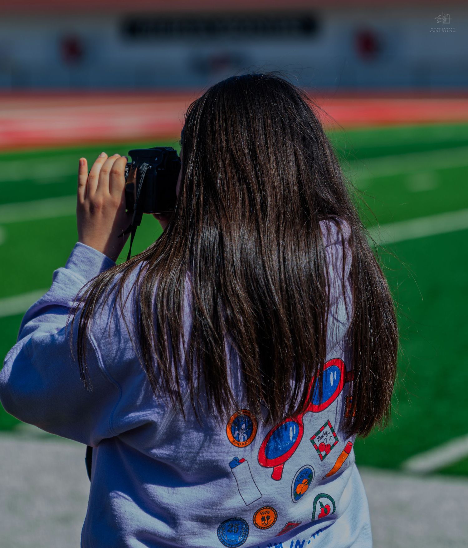 A girl with long dark hair photographs a green football field from the sidelines, wearing a colorful graphic sweatshirt.