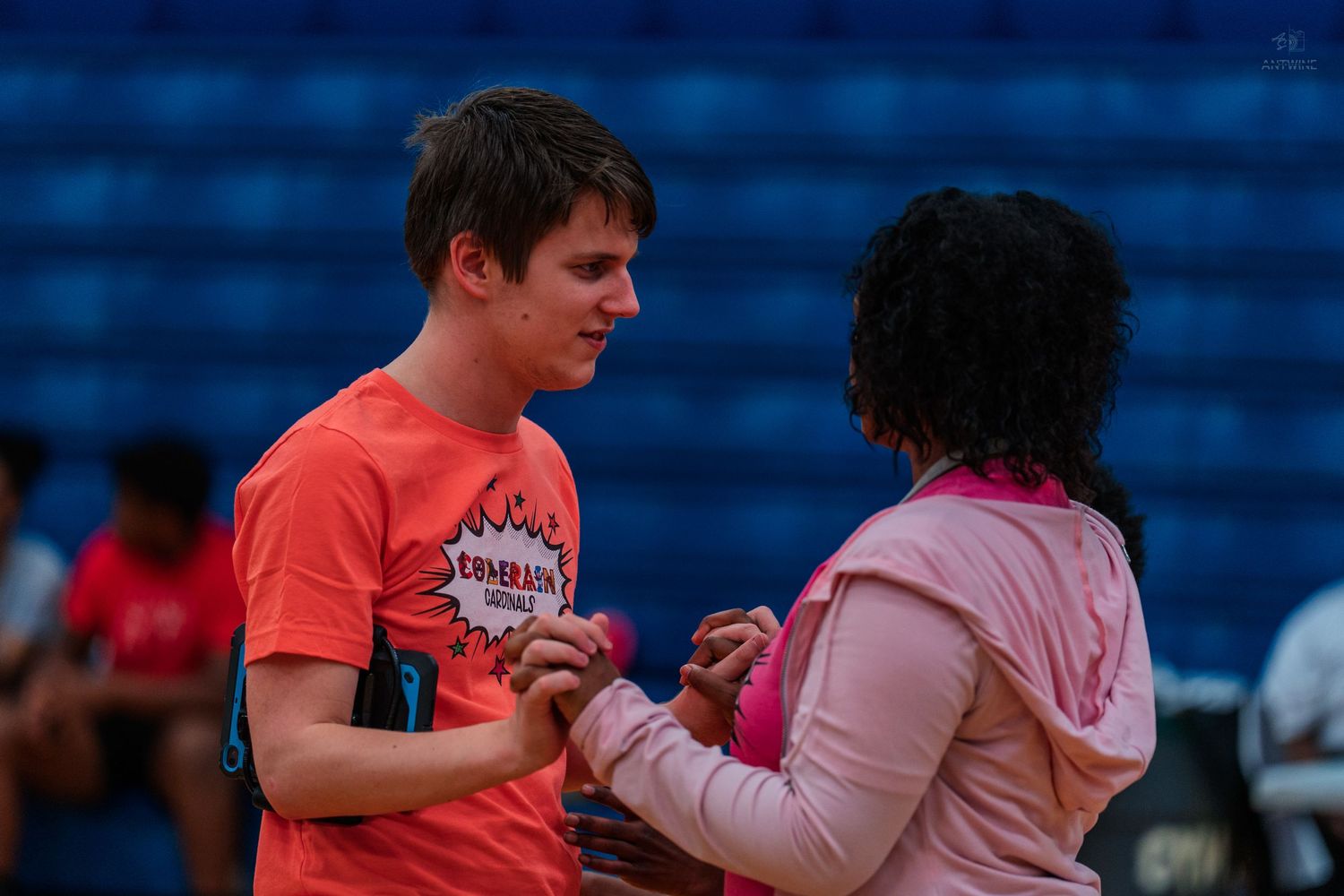 Two teens in athletic wear hold hands at an indoor sports event, with blue bleachers in the background.