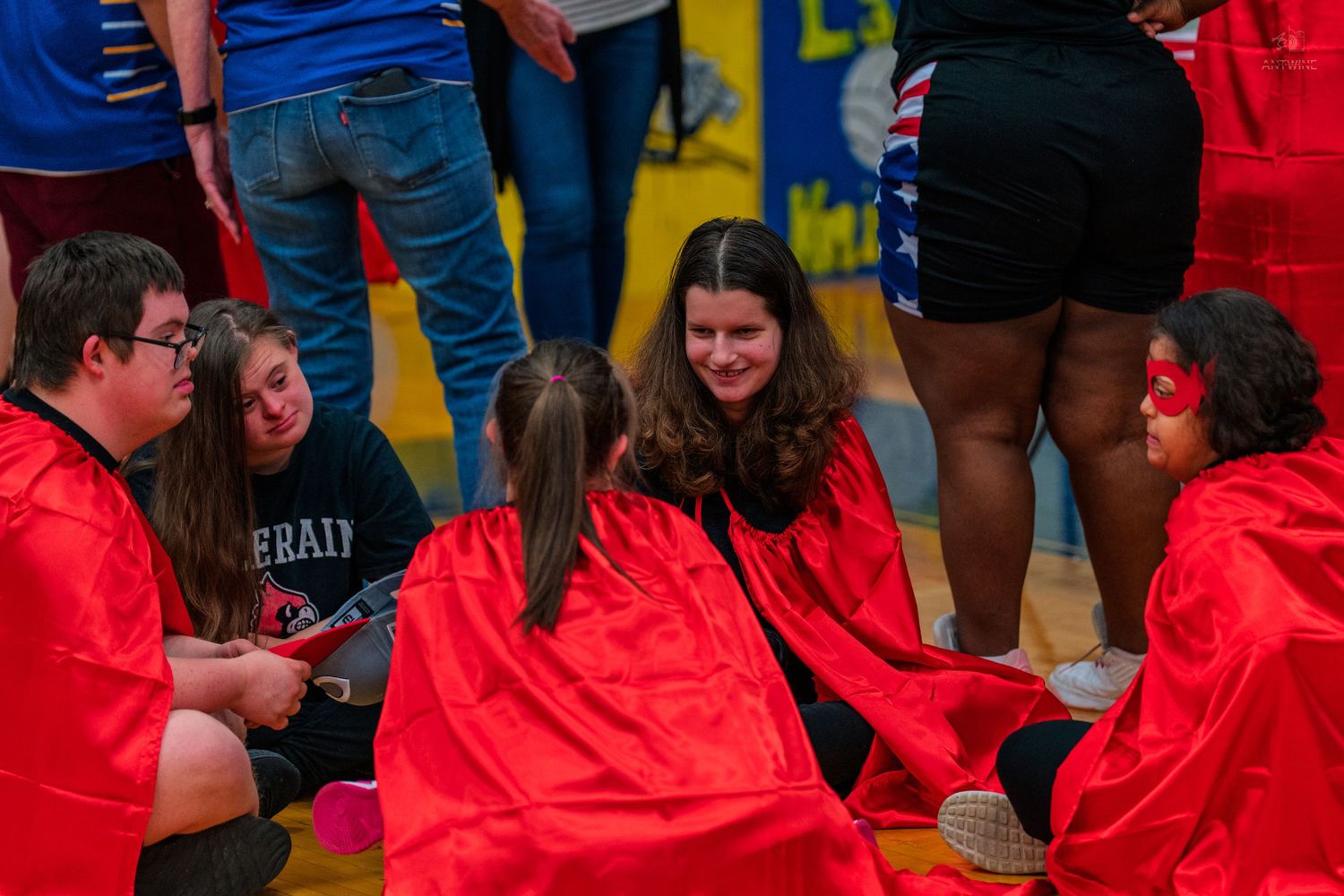 Students wearing red capes sit together on a gym floor during a school event, smiling and engaging in conversation.