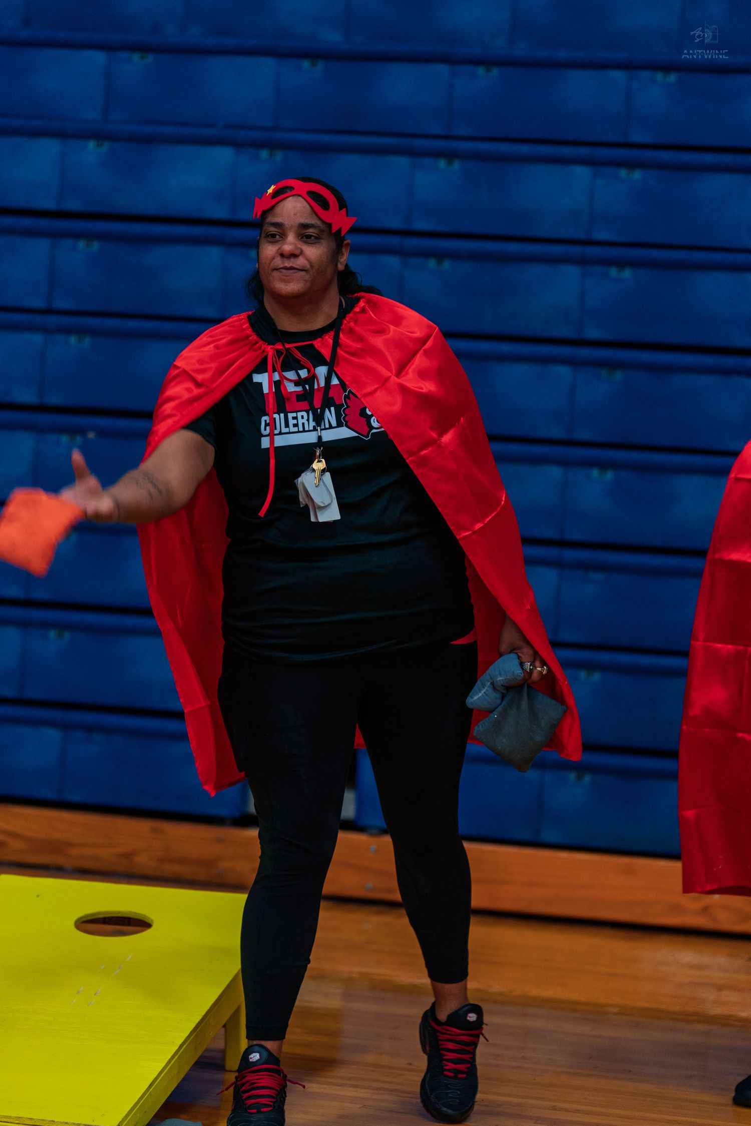 Woman in red cape and headband plays cornhole in a gymnasium, wearing a black outfit with sneakers.