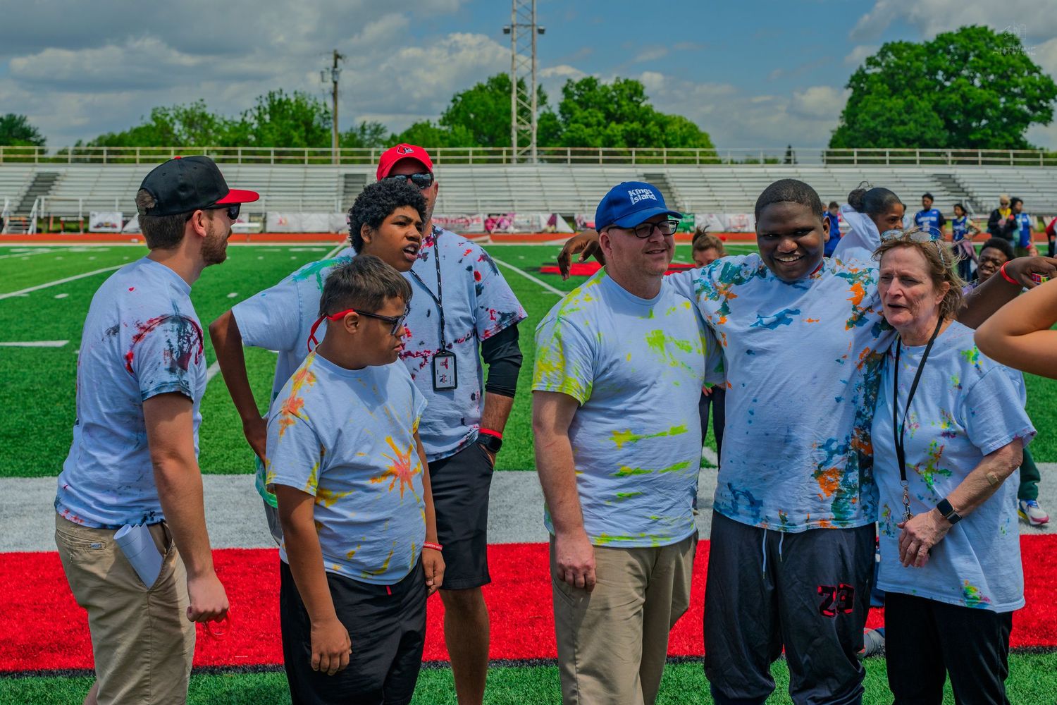 Group of adults in matching tie-dye shirts socializing on a football field during an outdoor sports event.