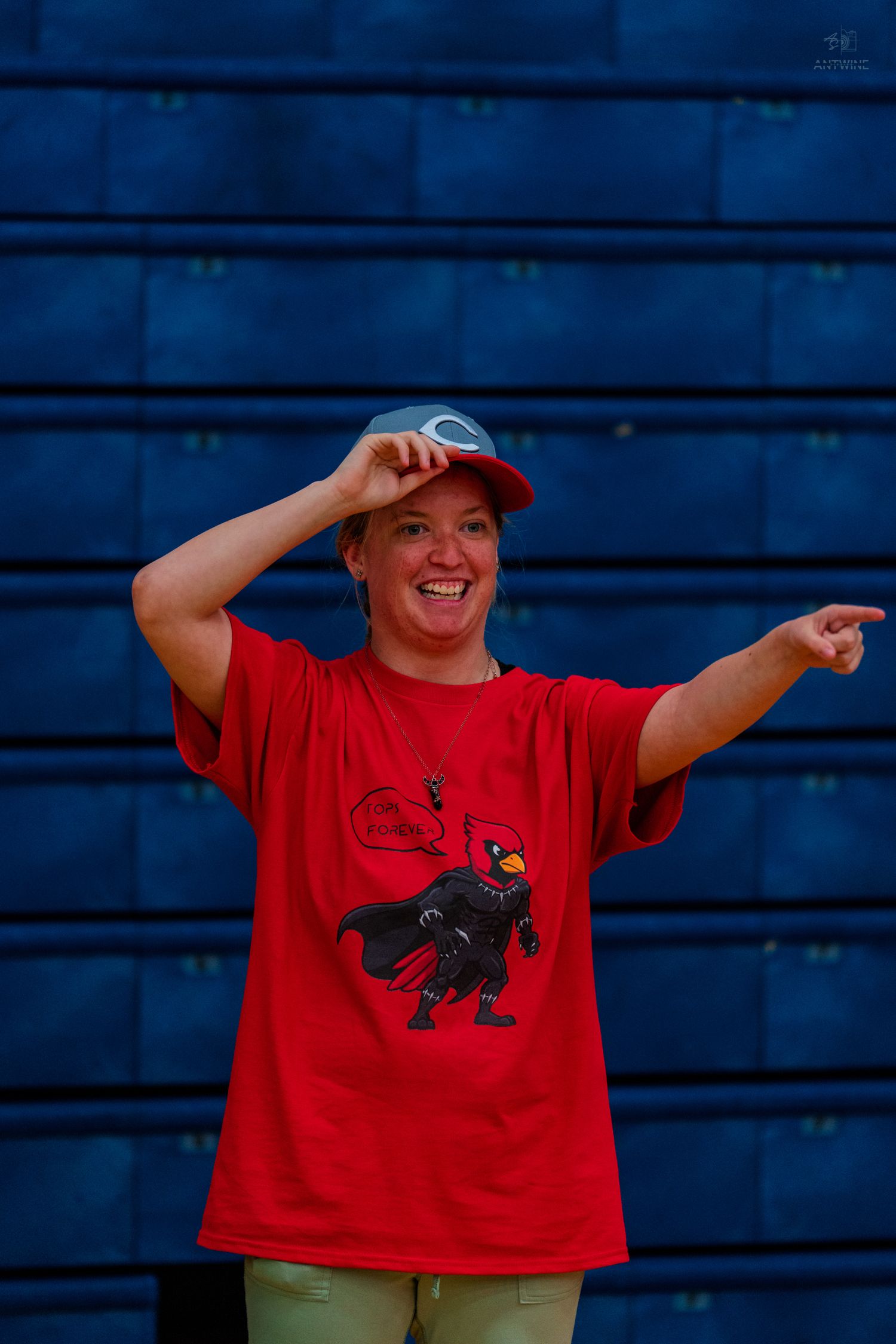Woman in red cardinal mascot t-shirt and cap smiling and pointing, standing in front of blue bleachers.