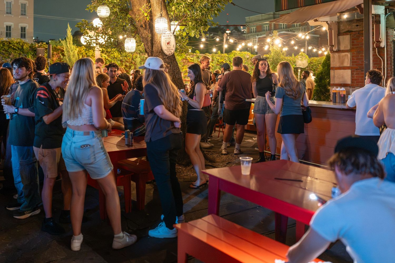 Young adults socializing at a lively outdoor bar patio with string lights at night.