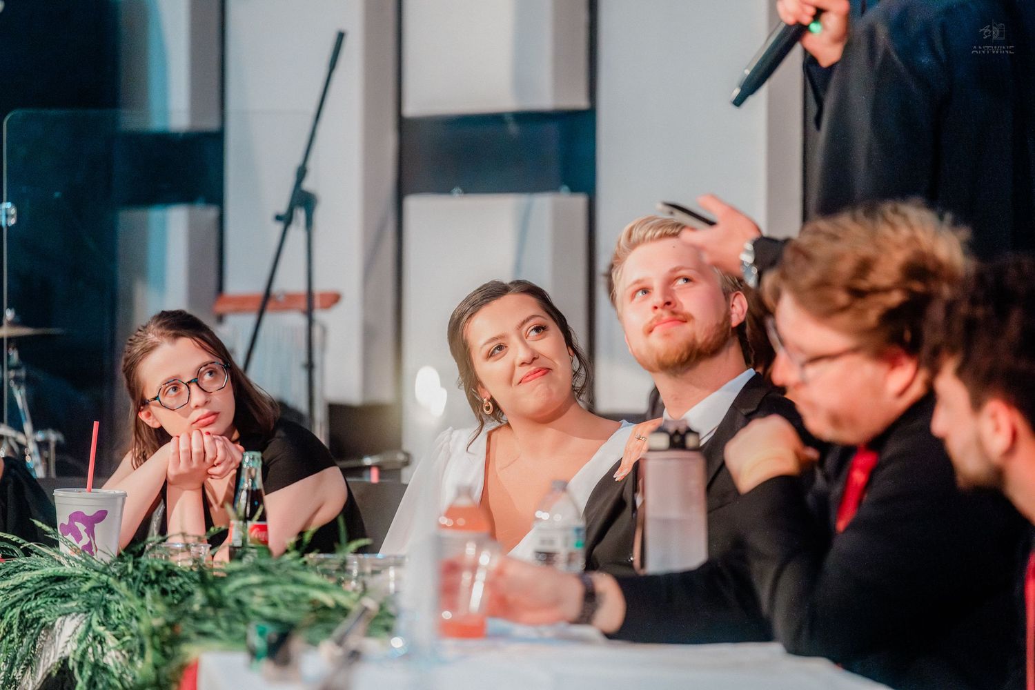 Wedding guests seated at a decorated table during a reception, with a man having his hair touched by someone standing behind.