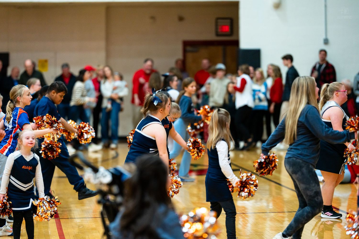 Mixed group of cheerleaders in navy and black uniforms dance with pom-poms in a crowded gymnasium.