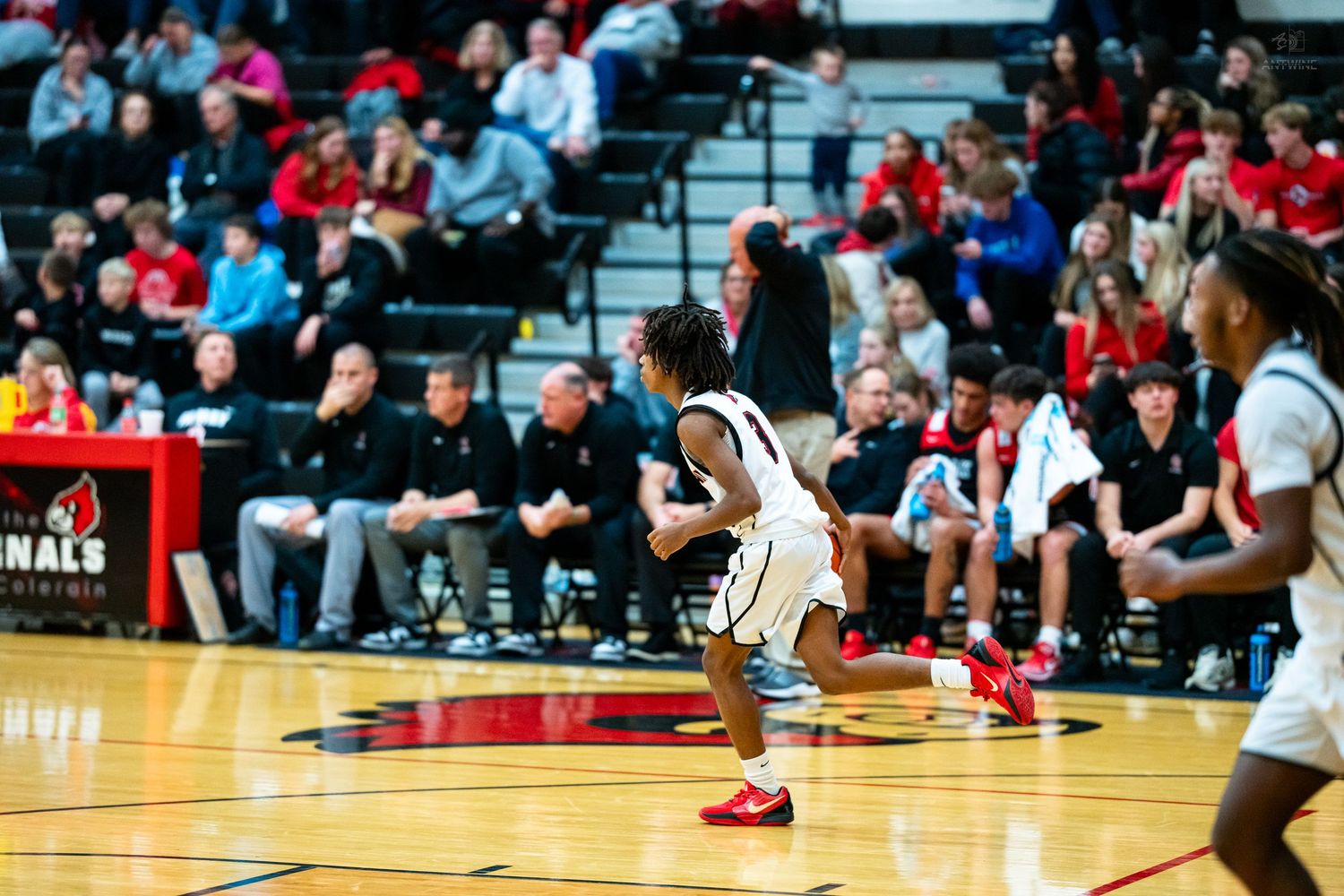 Basketball player in red uniform celebrates on the court during a high school game with teammates and fans nearby.
