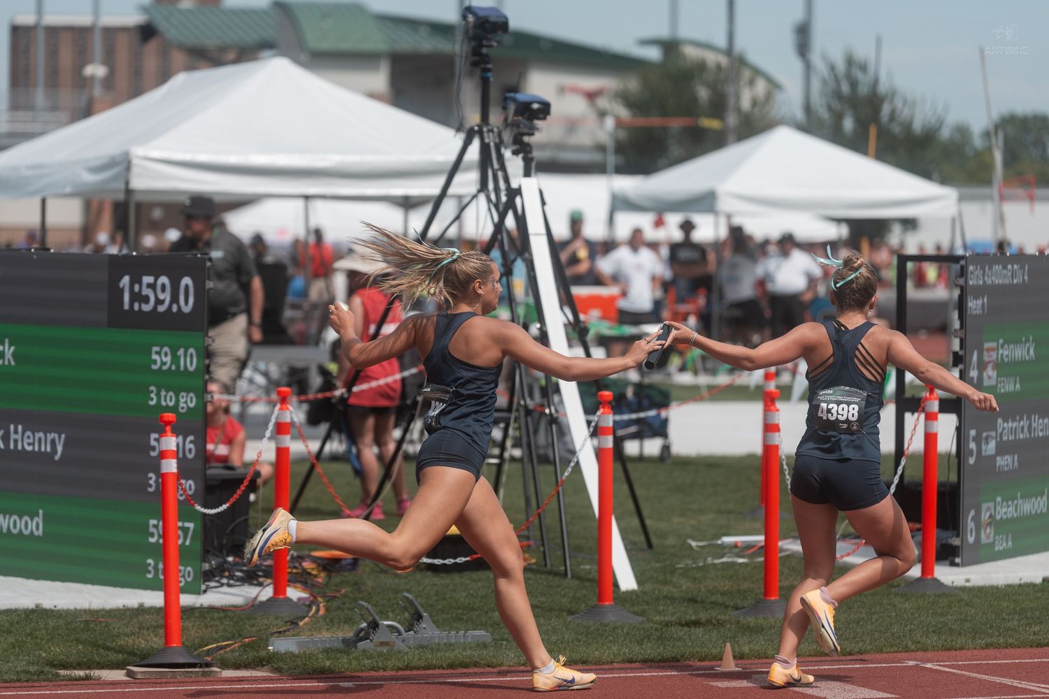 Two female relay runners execute a baton exchange during a high school track and field championship race.