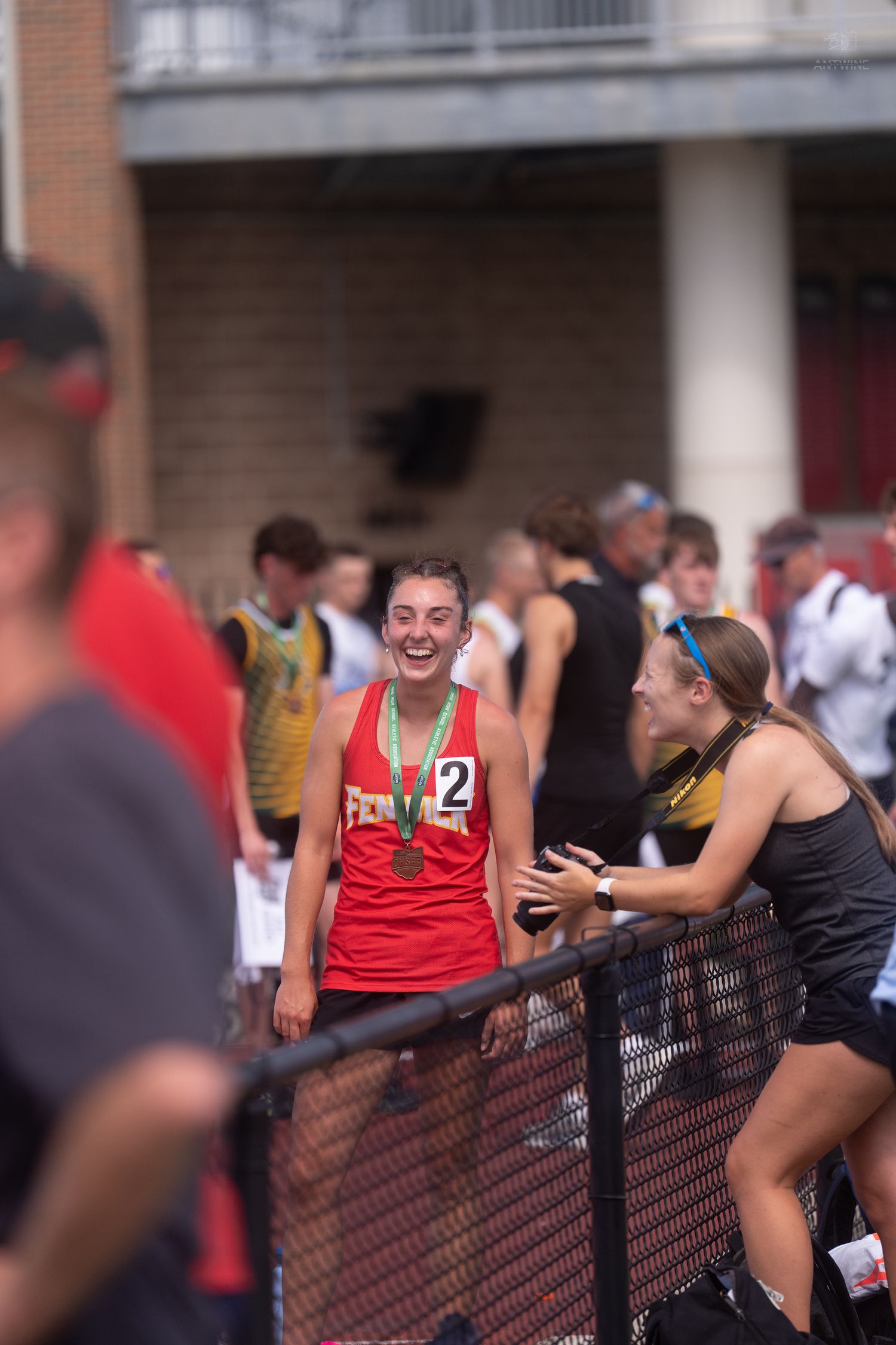Smiling female athlete wearing number 2 jersey and medal celebrates at track meet.