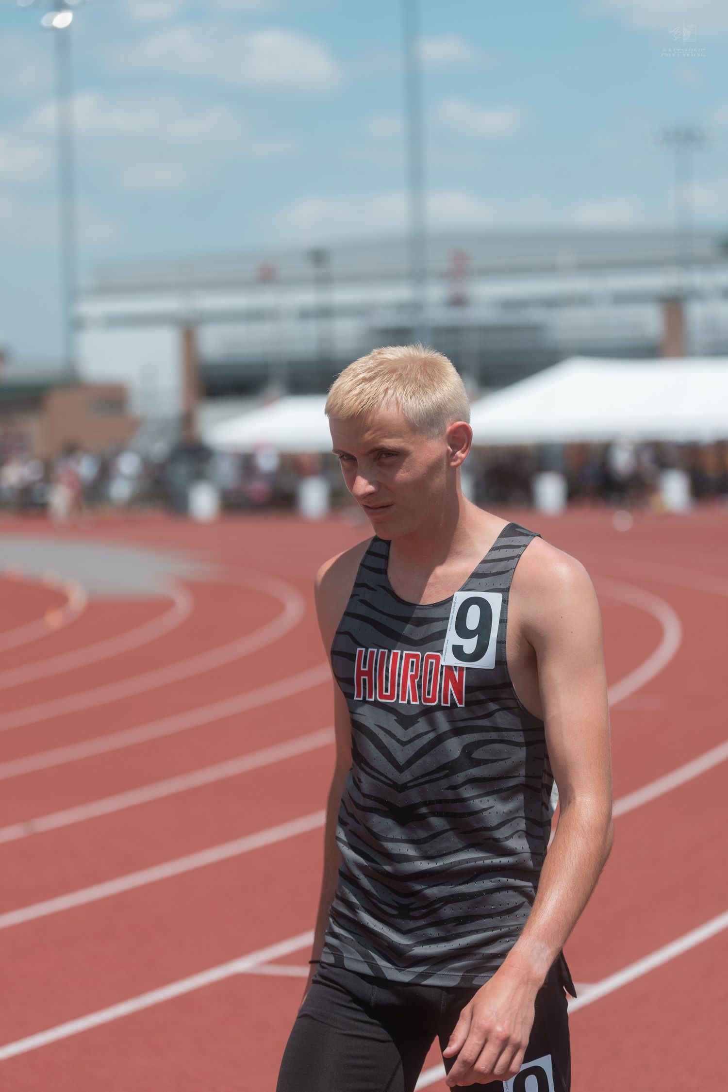 A blonde male track athlete wearing a Huron number 9 jersey stands on a red track on a sunny day.