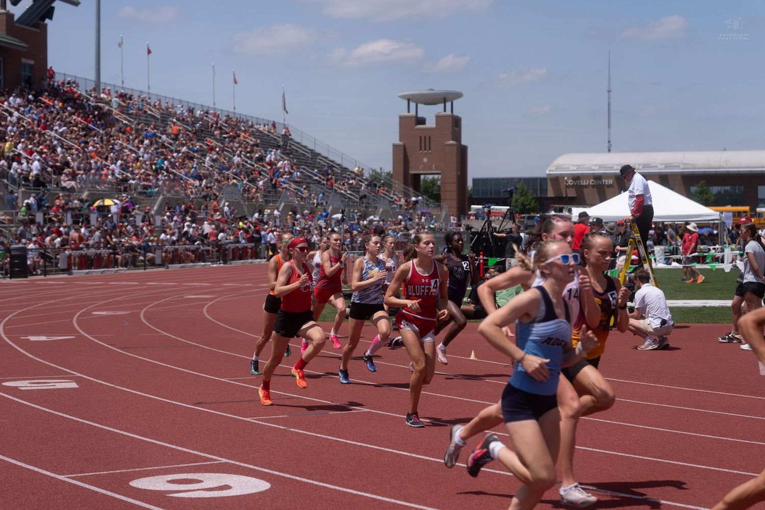 High school girls compete in a track and field race at a crowded outdoor stadium on a sunny day.