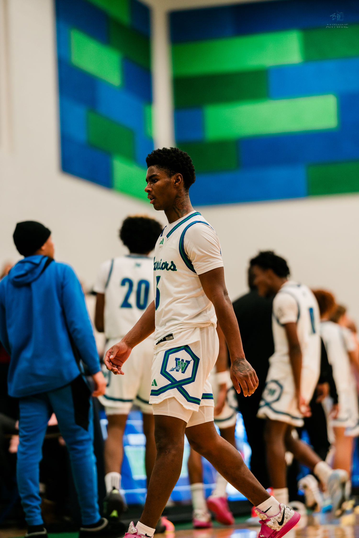 A Warriors basketball player walks near the sideline during a high school game with teammates in the background.