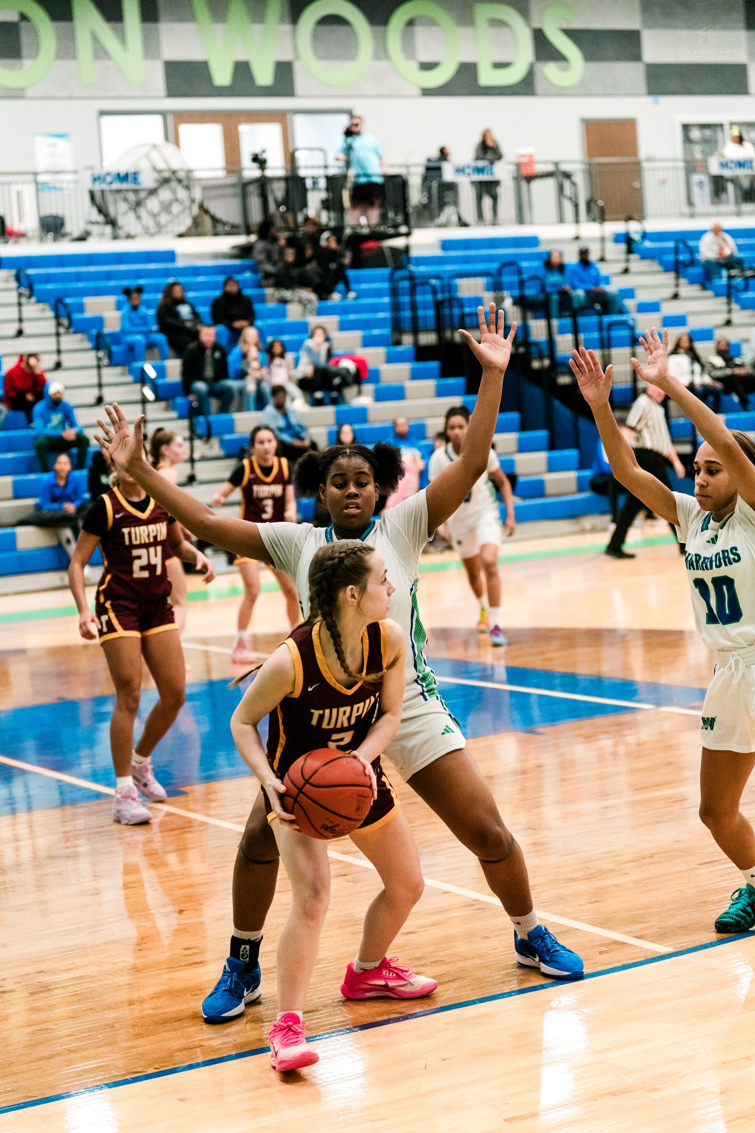 Turpin girls basketball player in maroon holds ball while surrounded by Warriors defenders with hands raised at Mason Woods gym.