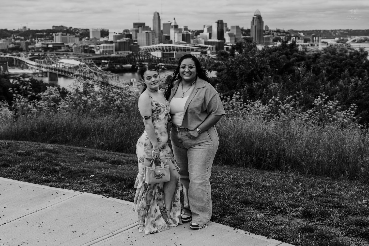 Two women posing together at scenic Cincinnati overlook with city skyline in black and white.