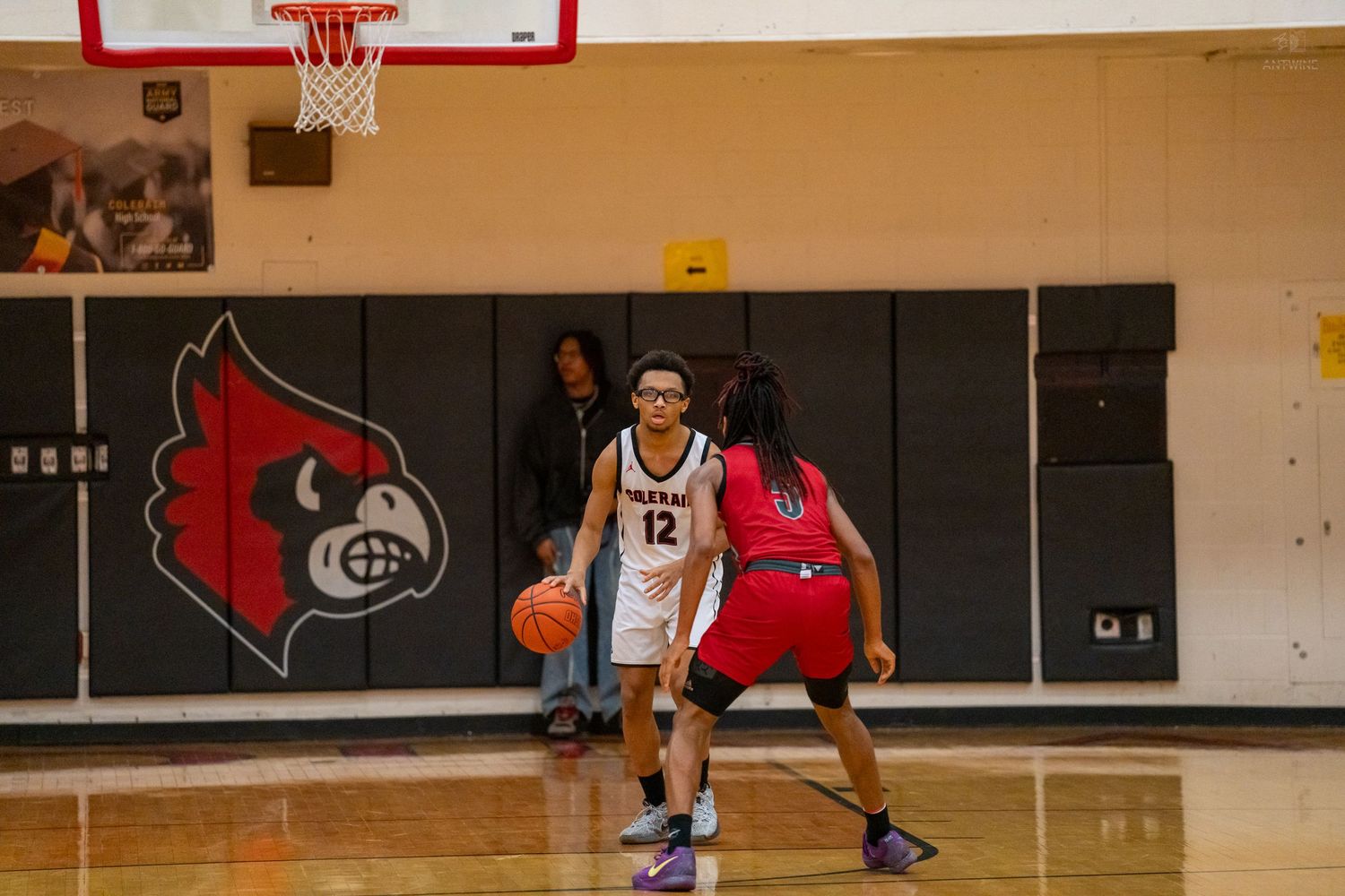 High school basketball player number 12 in white dribbles against red-uniformed defender near Cardinals mascot wall.