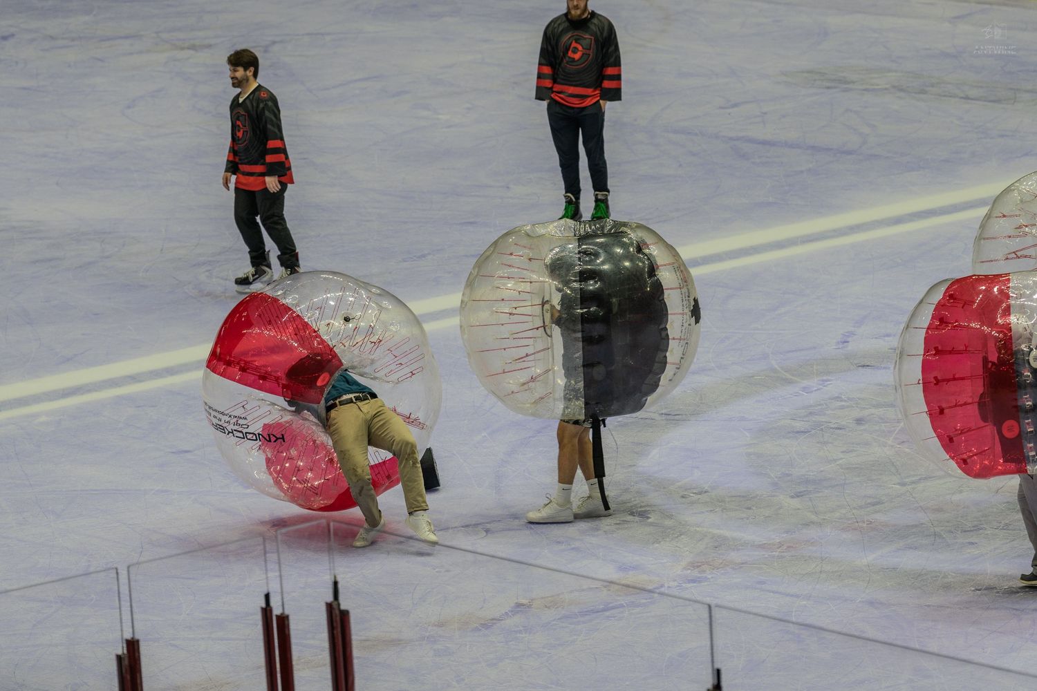 A player bends over while inside a bubble soccer zorb ball on an ice rink with hockey players watching.