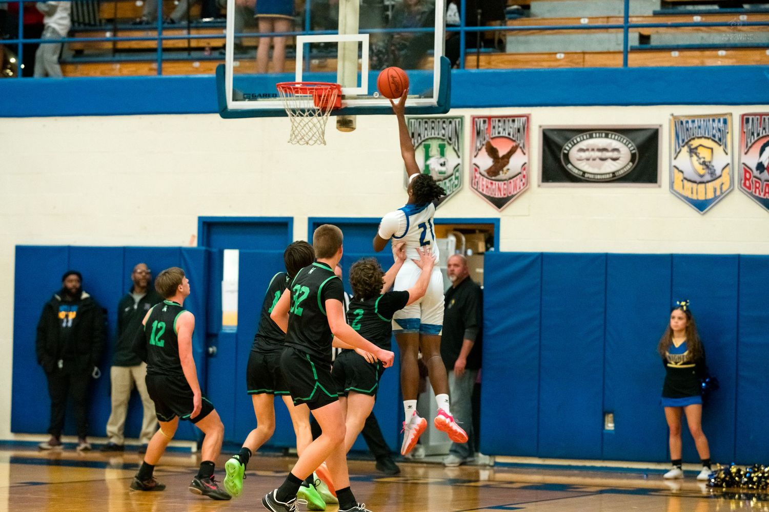 Tall player goes up for basket against green-uniformed defenders near hoop.