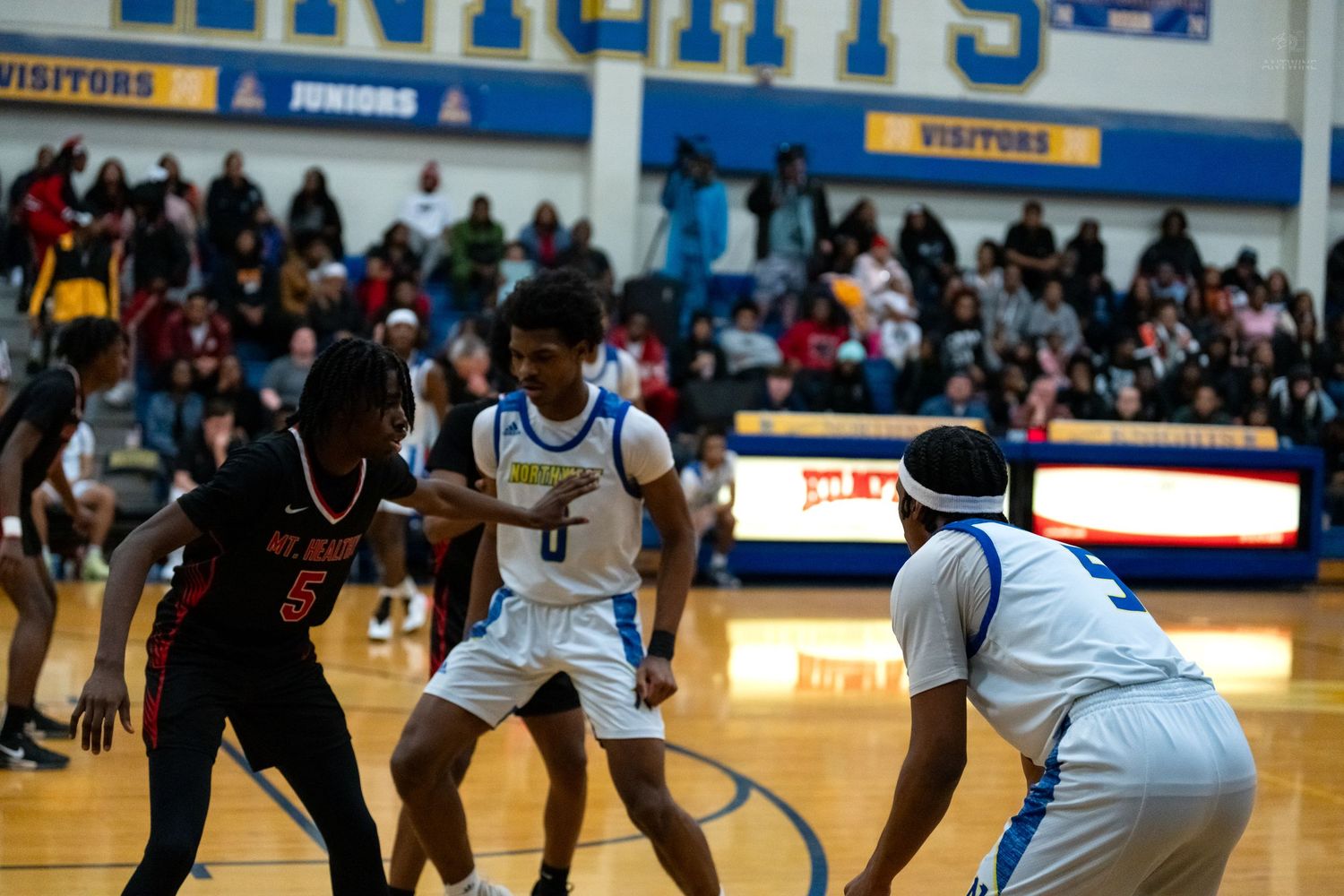 High school basketball players in blue and red uniforms compete in a defensive play during a gymnasium game.