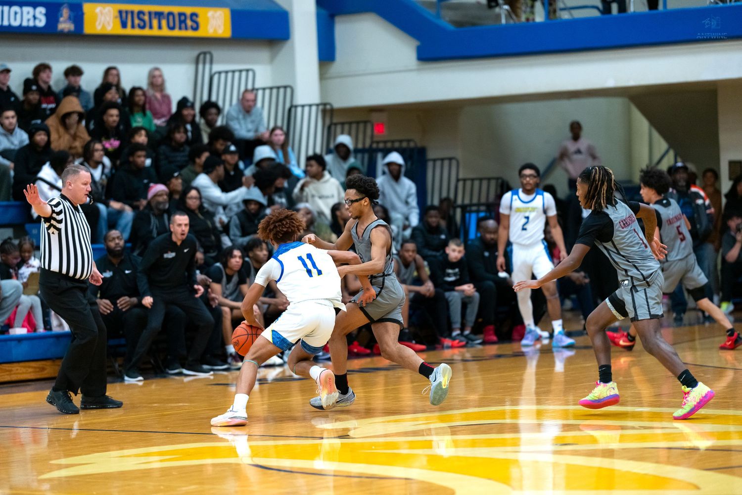 High school basketball player number 11 in white dribbles past defenders during a crowded gymnasium game.
