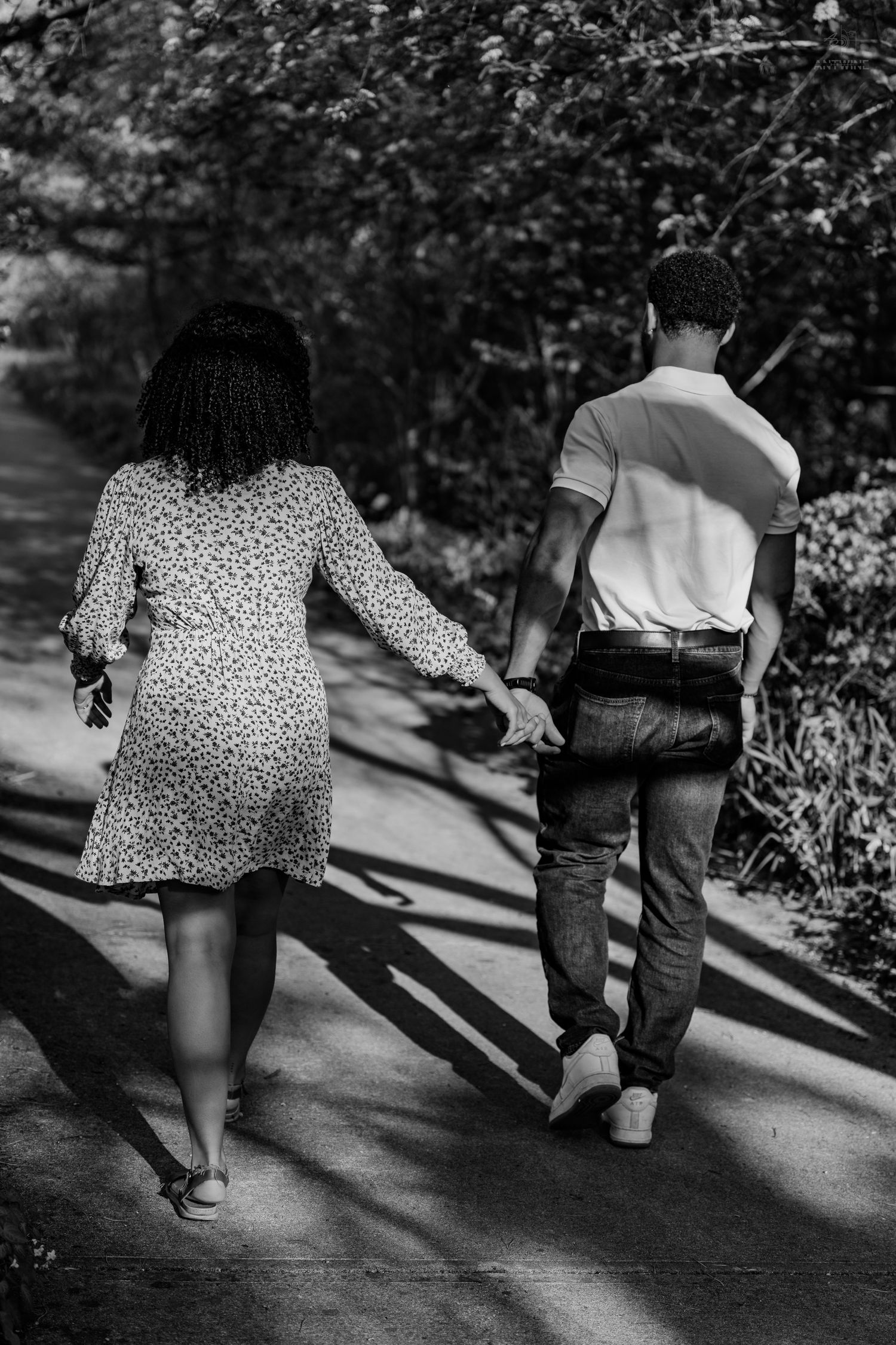 Black and white rear view of couple holding hands walking down a tree-lined garden path together.
