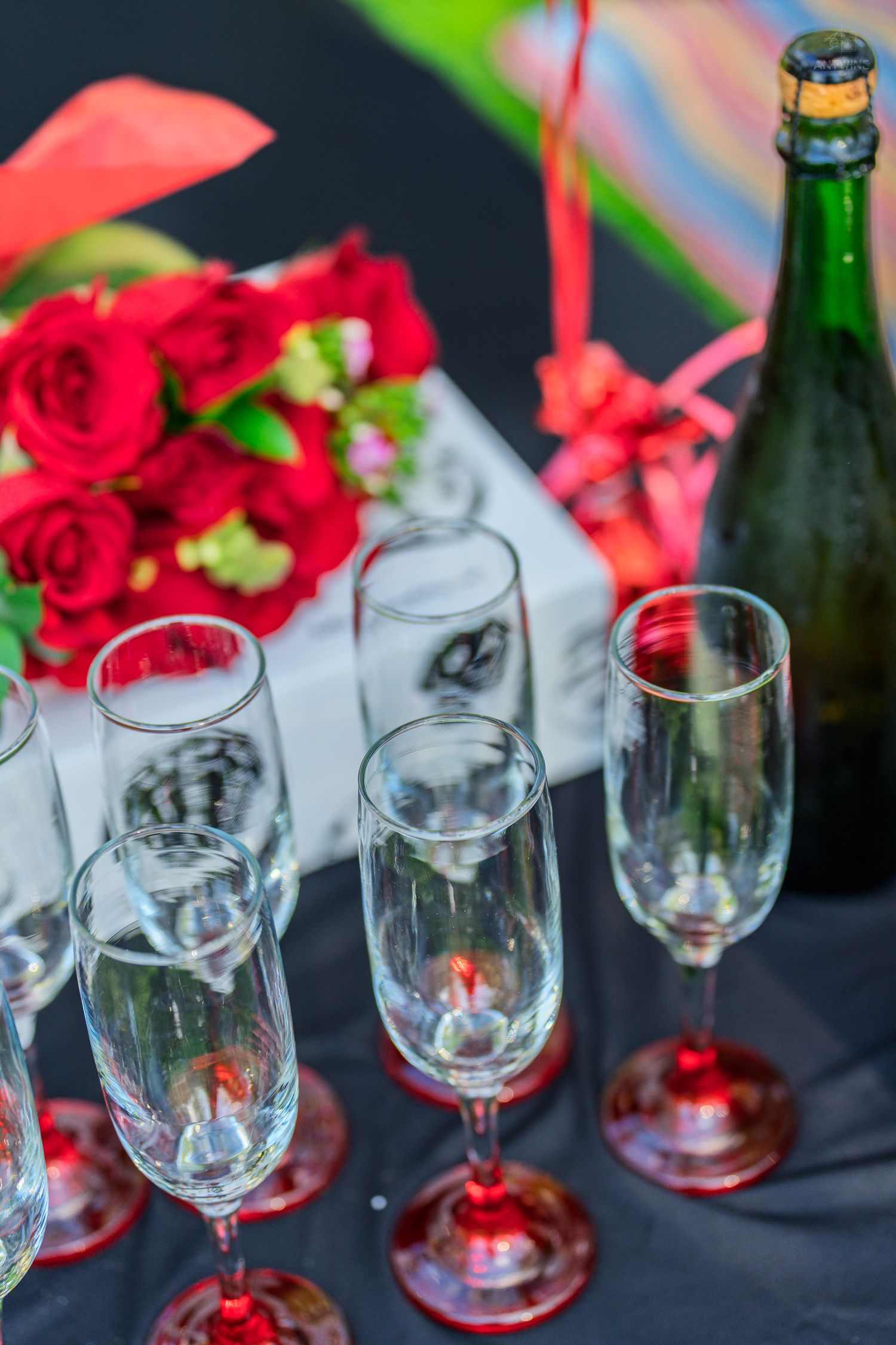 Champagne flutes with red bases beside a bouquet of red roses and a gift box on a black tablecloth.
