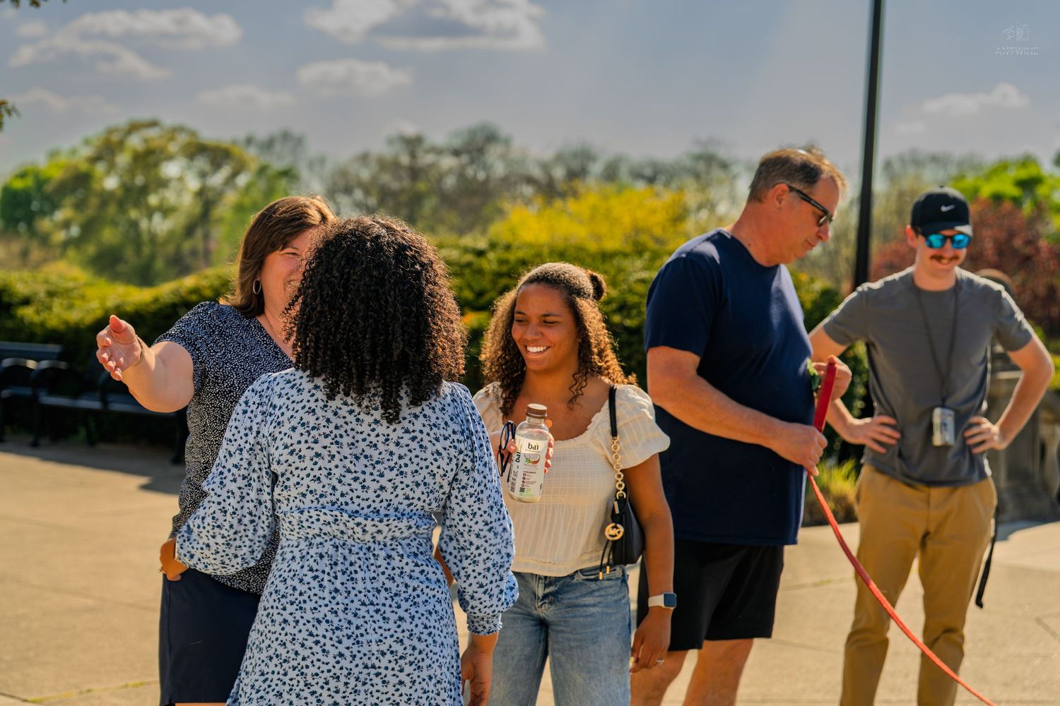 Diverse group of people socializing outdoors on a sunny day with trees in the background.