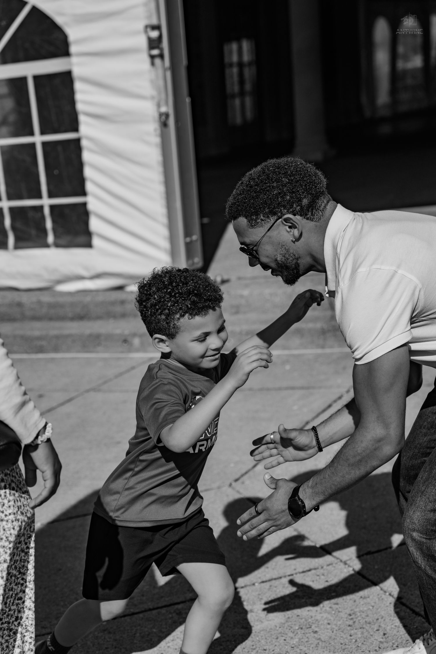 Black and white photo of a smiling young boy running to high-five a man outdoors near a white tent.