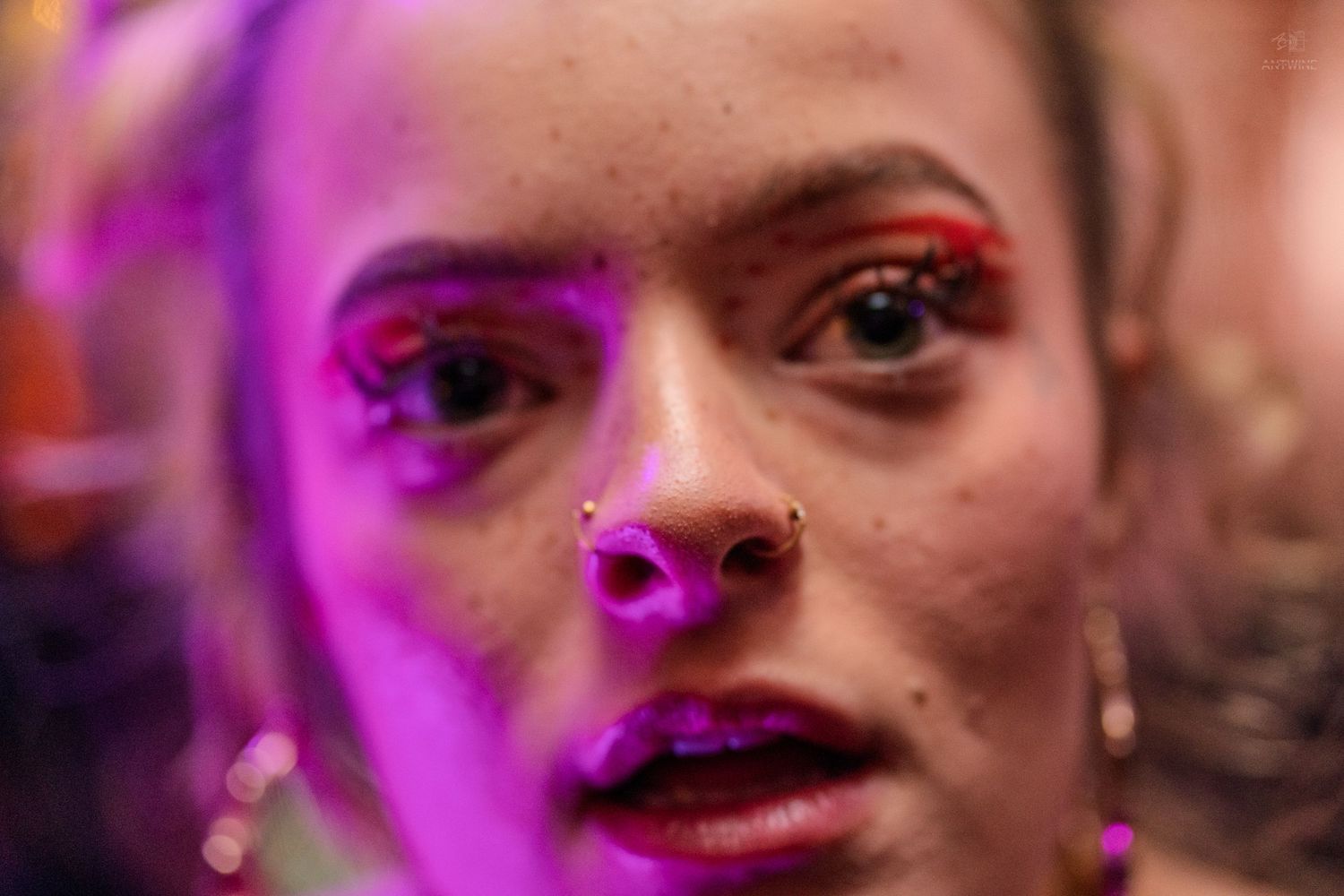 Close-up portrait of a woman with nose piercings and bold red eye makeup under pink neon light.
