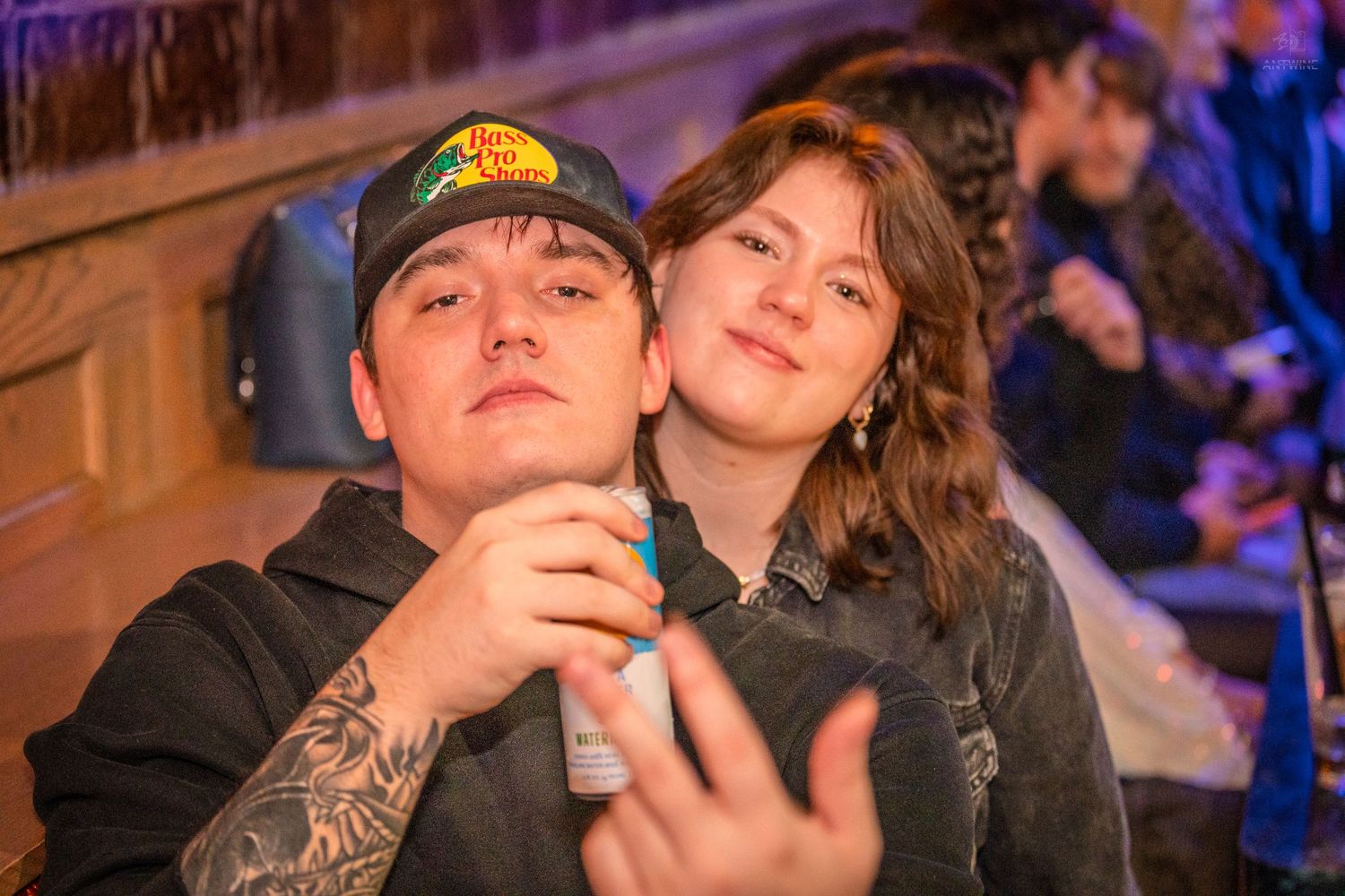 Young couple posing together at a bar, man holding a canned drink with tattoos visible.