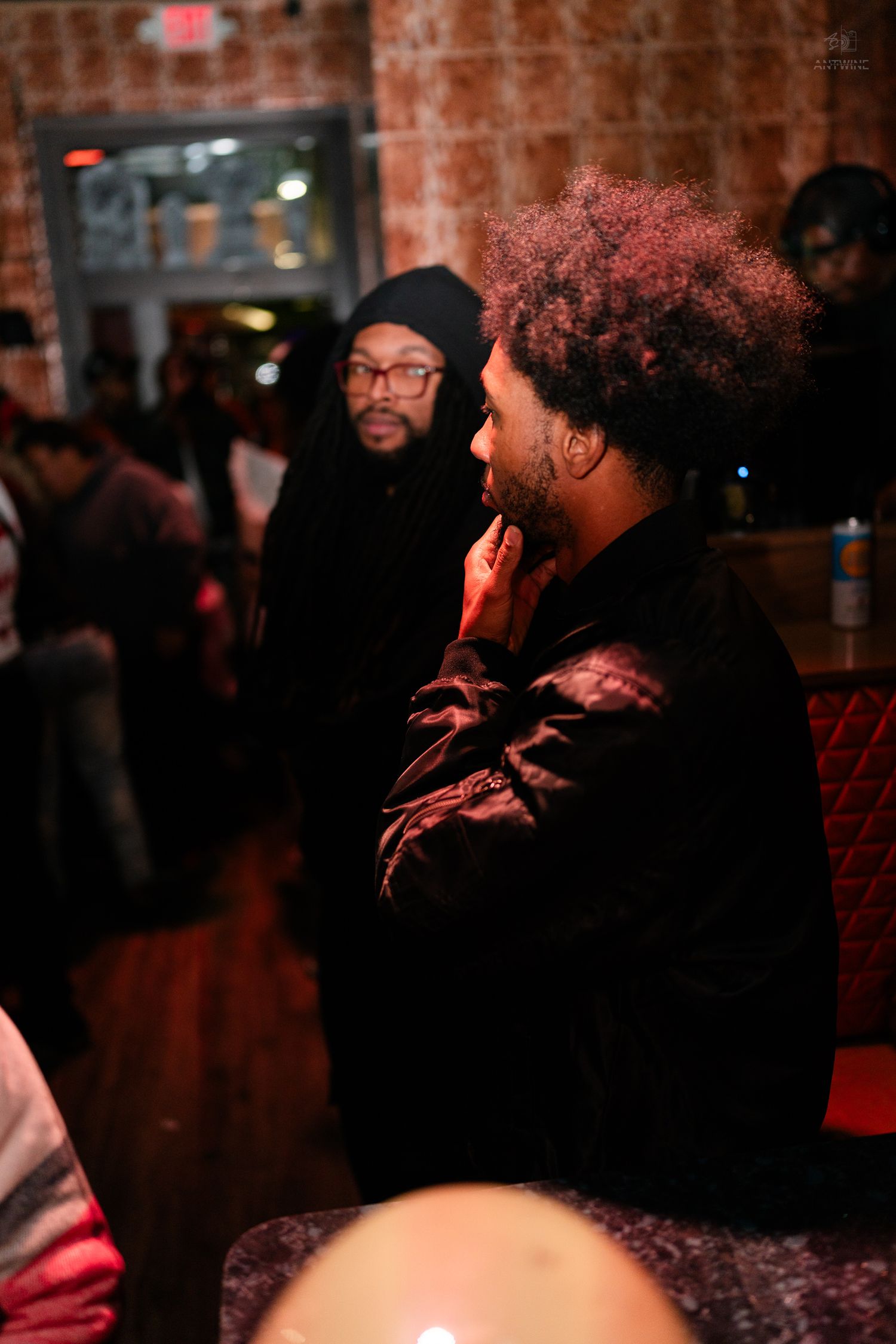 Two men having a conversation in a warmly lit bar, one with dreadlocks and glasses, another with curly hair.