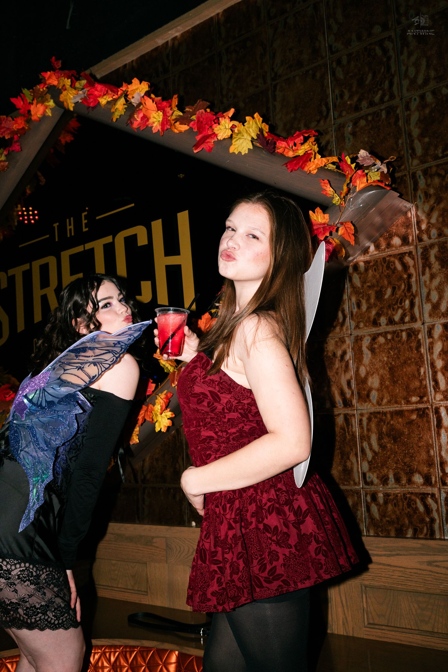 Two women in Halloween costumes pose at The Stretch bar decorated with autumn leaves.