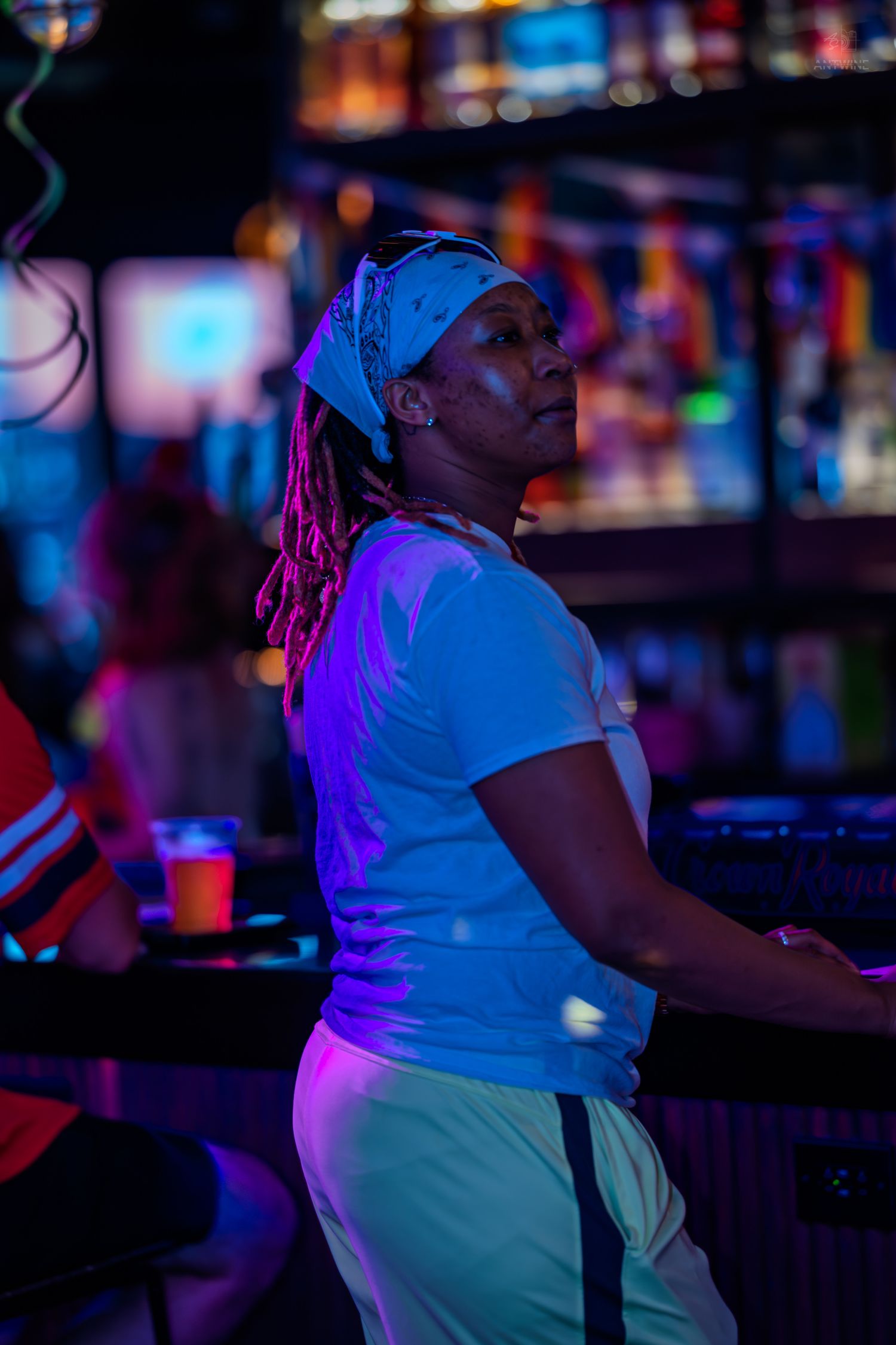 A Black woman with dreadlocks and a bandana poses in a neon-lit bar wearing a white tee.